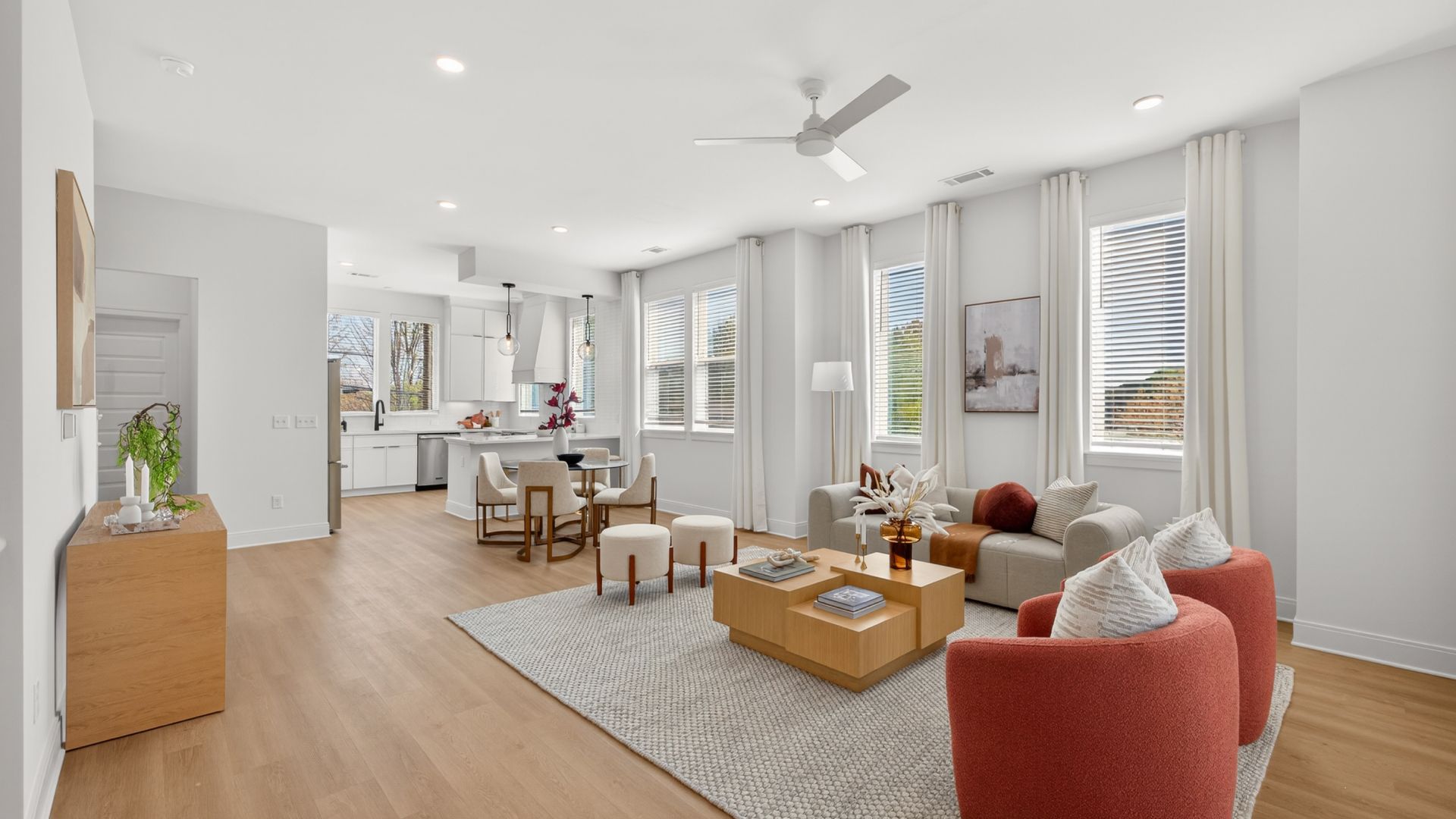 Bright open-plan living room and kitchen with white walls, light wood floors, a ceiling fan, and large windows with white curtains. Beige sofa, red curved chairs and wood coffee tables on a gray rug.