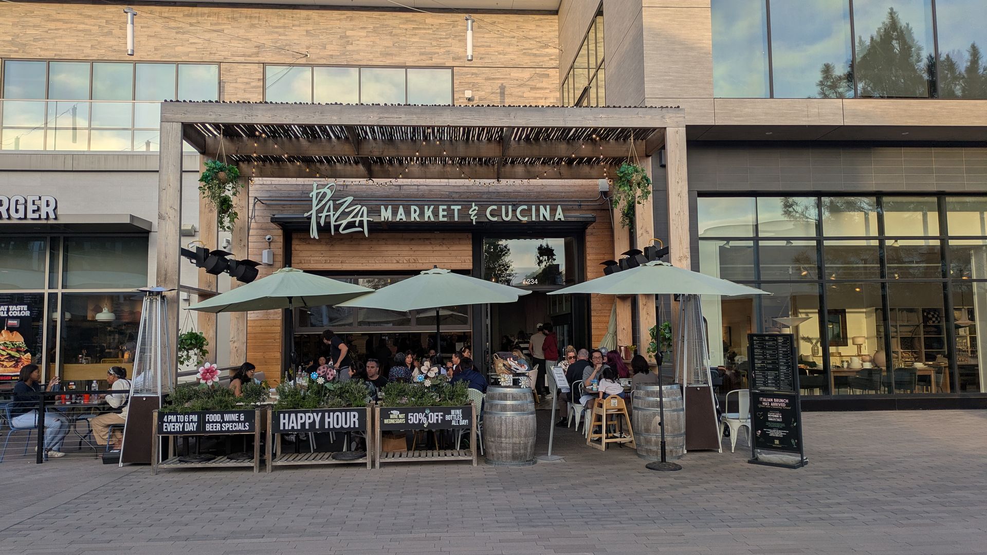 Modern shopping plaza with a restaurant sign reading "Piaza Market & Cucina" under a wooden pergola and string lights; outdoor seating with umbrellas, large glass windows reflecting trees and blue sky.