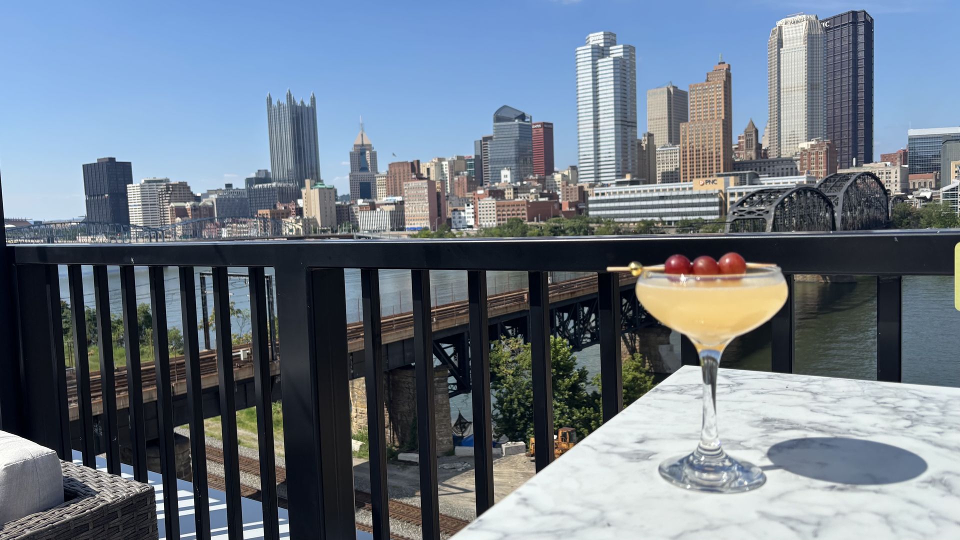 Cocktail with red grape garnish on a marble table overlooking Pittsburgh skyline and Allegheny River on a clear sunny day.