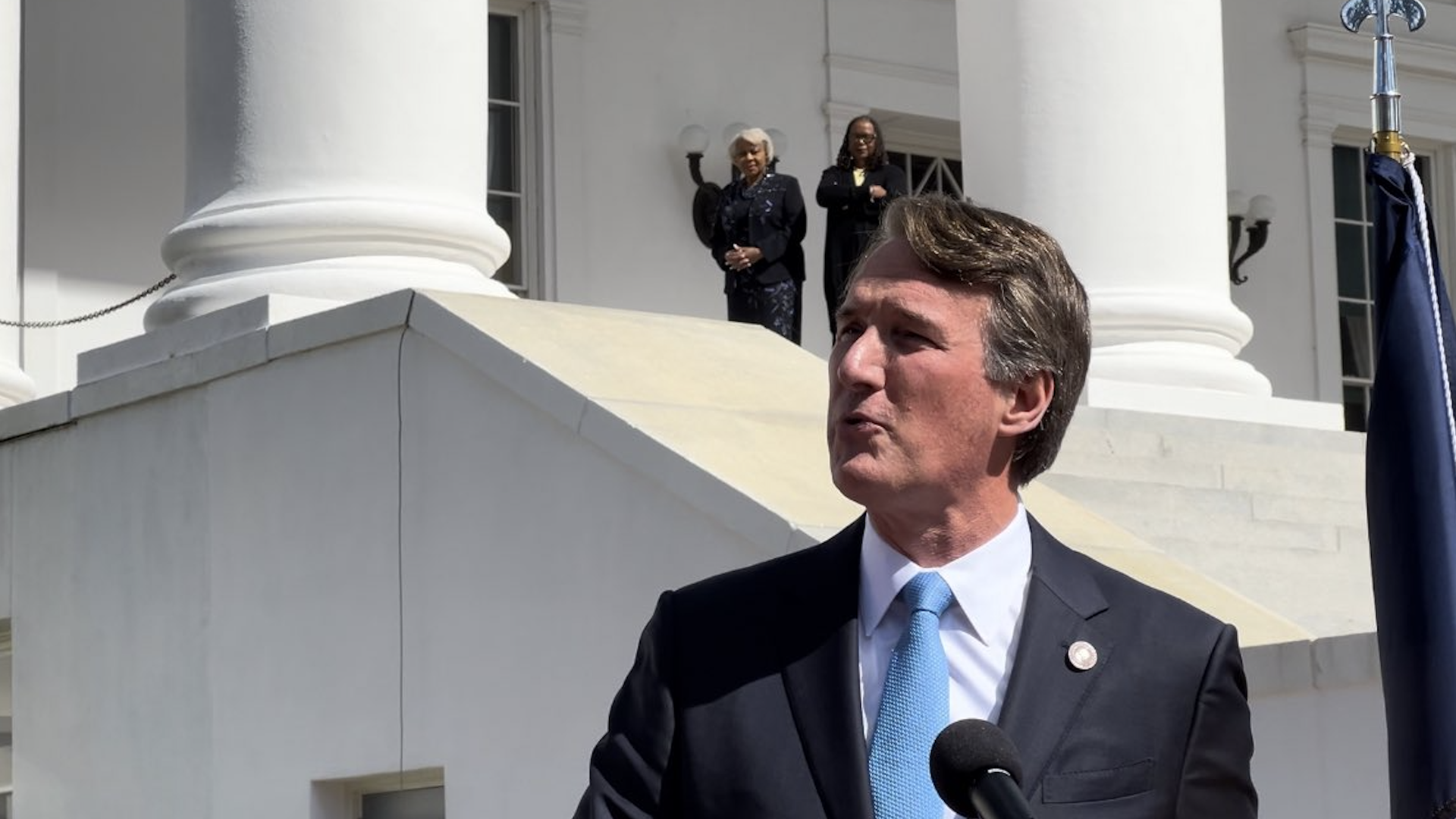 Glenn Youngkin speaks before a microphone, with Sen. Louise Lucas seen in the background