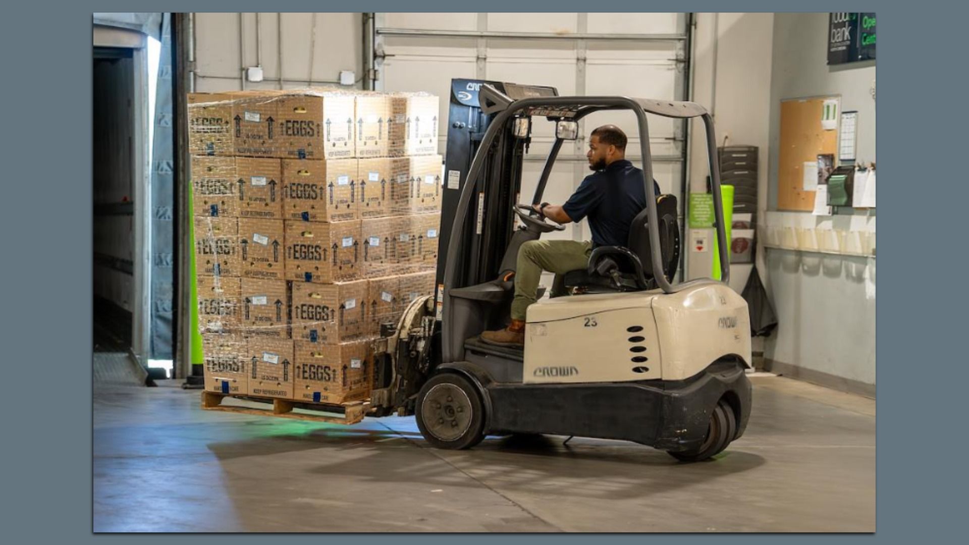 A warehouse worker drives a small forklift beside tall pallets of cardboard boxes labeled "EGGS," moving them toward a loading dock; background shows a doorway and office boards.
