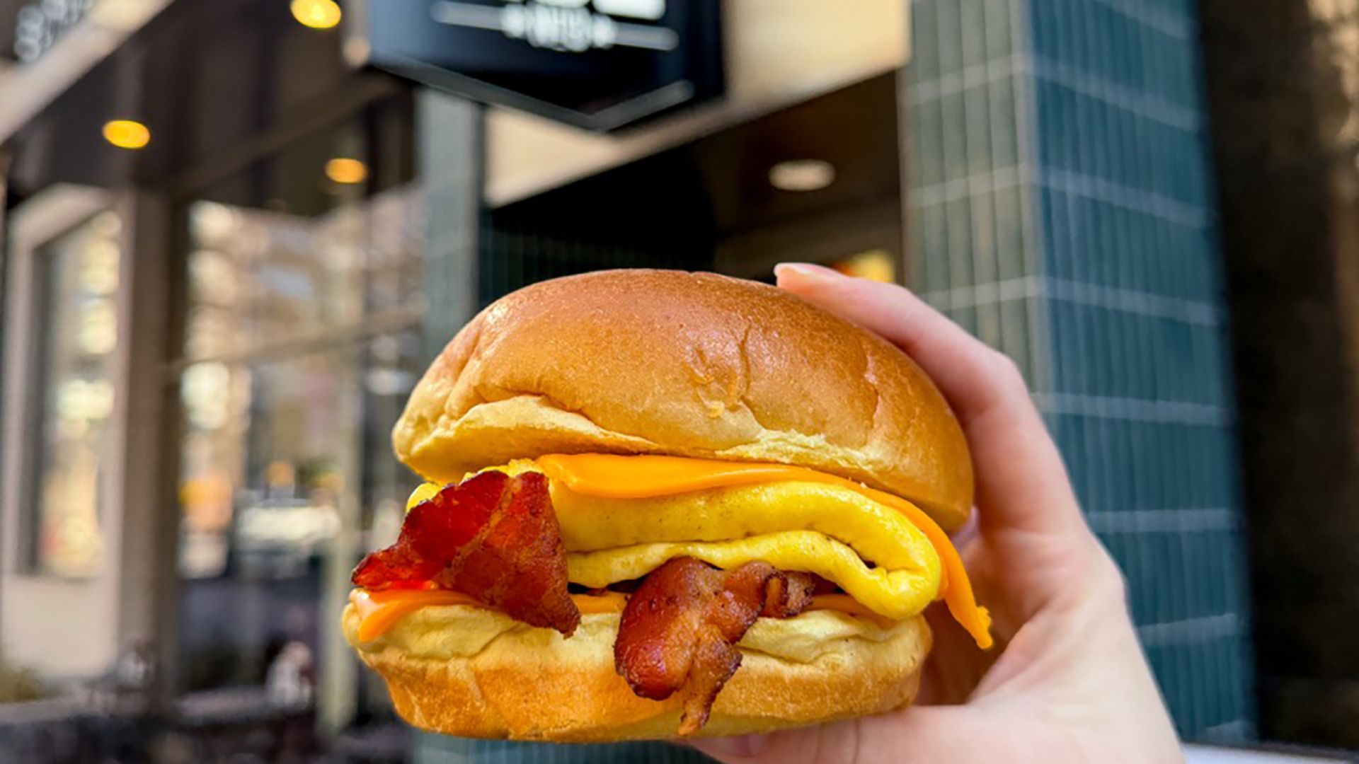 Hand holding a breakfast sandwich with bacon, folded yellow egg, and cheddar cheese in a soft bun outside a cafe with a black and white "ACE" sign.