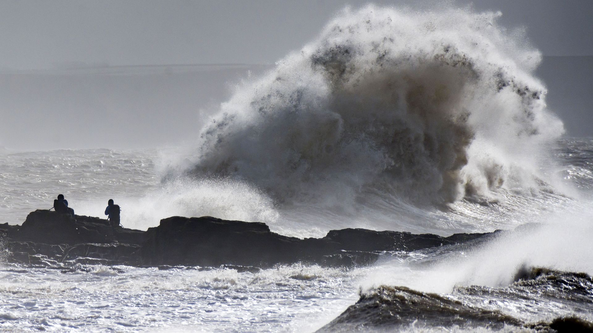 Waves crash near the harbour wall as people look on