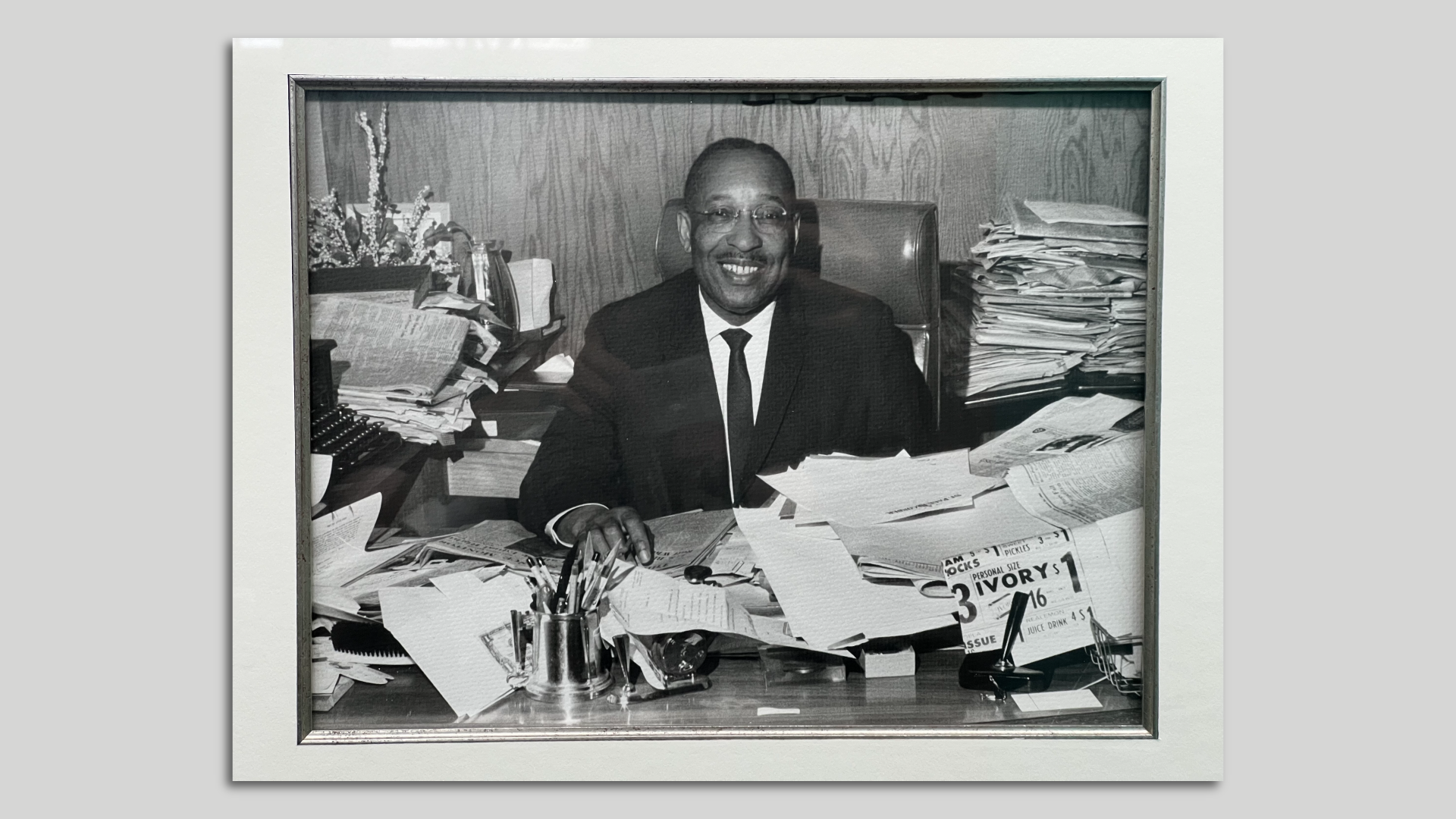 A black-and-white photo of a man with dark skin in a suit and tie sitting and smiling at a desk strewn with newsprint.