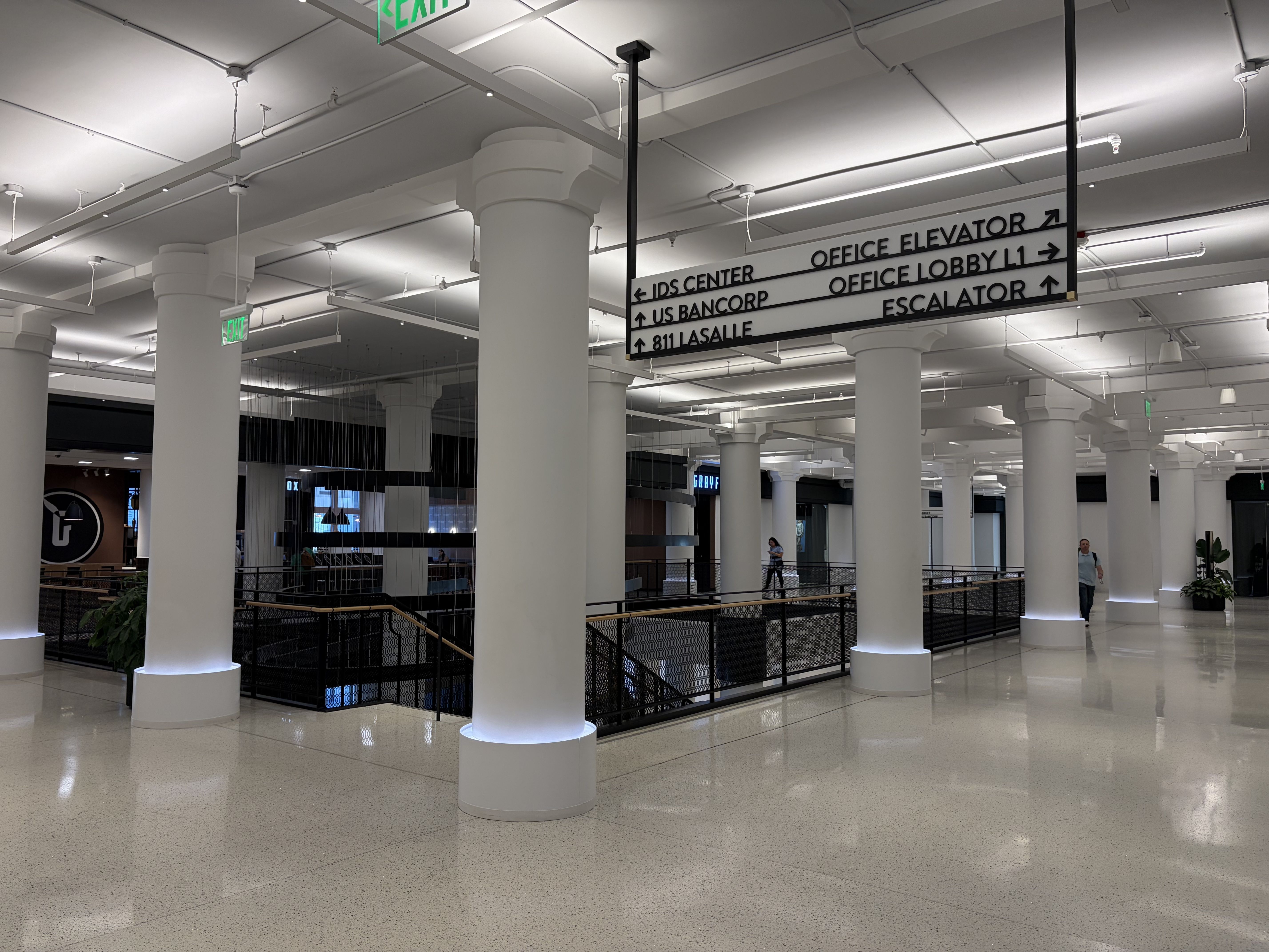 Indoor lobby with tall white columns, bright ceiling lights, and a large black-and-white directory sign listing offices. Green exit signs, glass railings, and a few people in the distance.