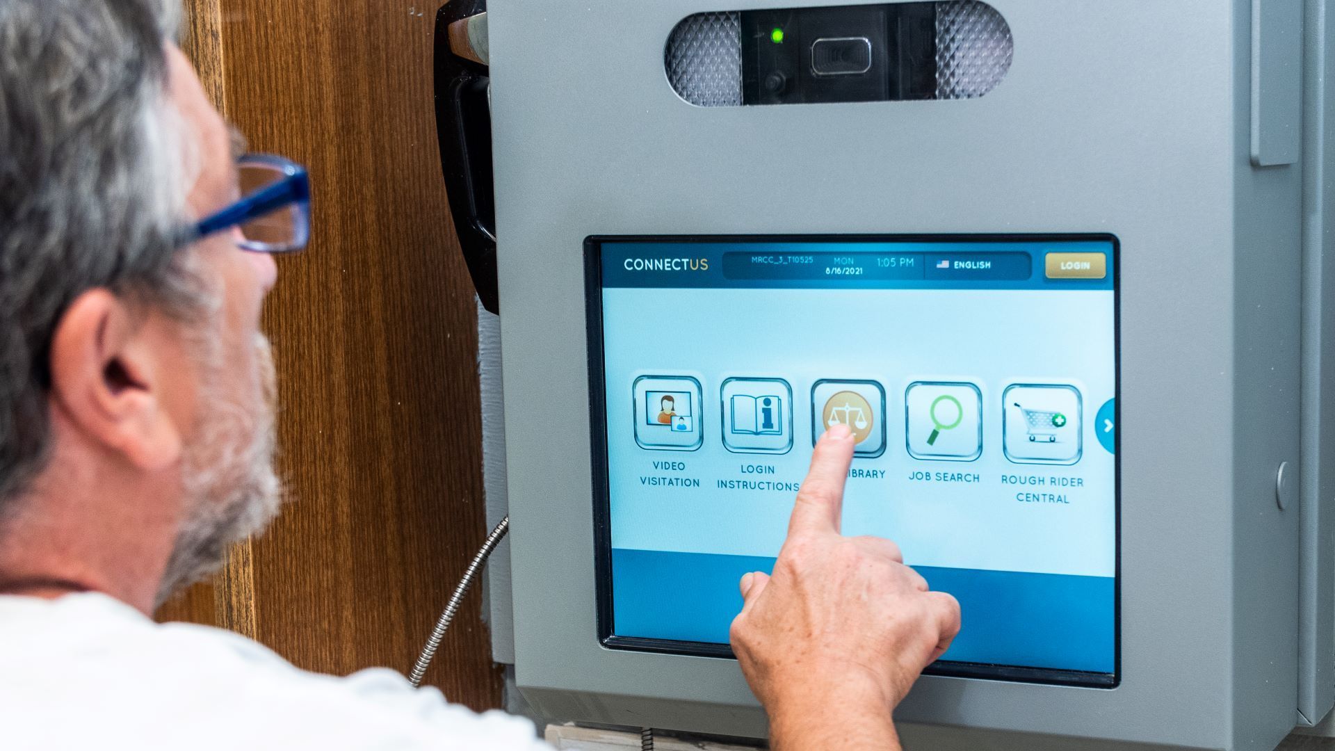 A person interacts with a digital kiosk providing services like video visitation, legal resources, and job searches, within a correctional facility.
