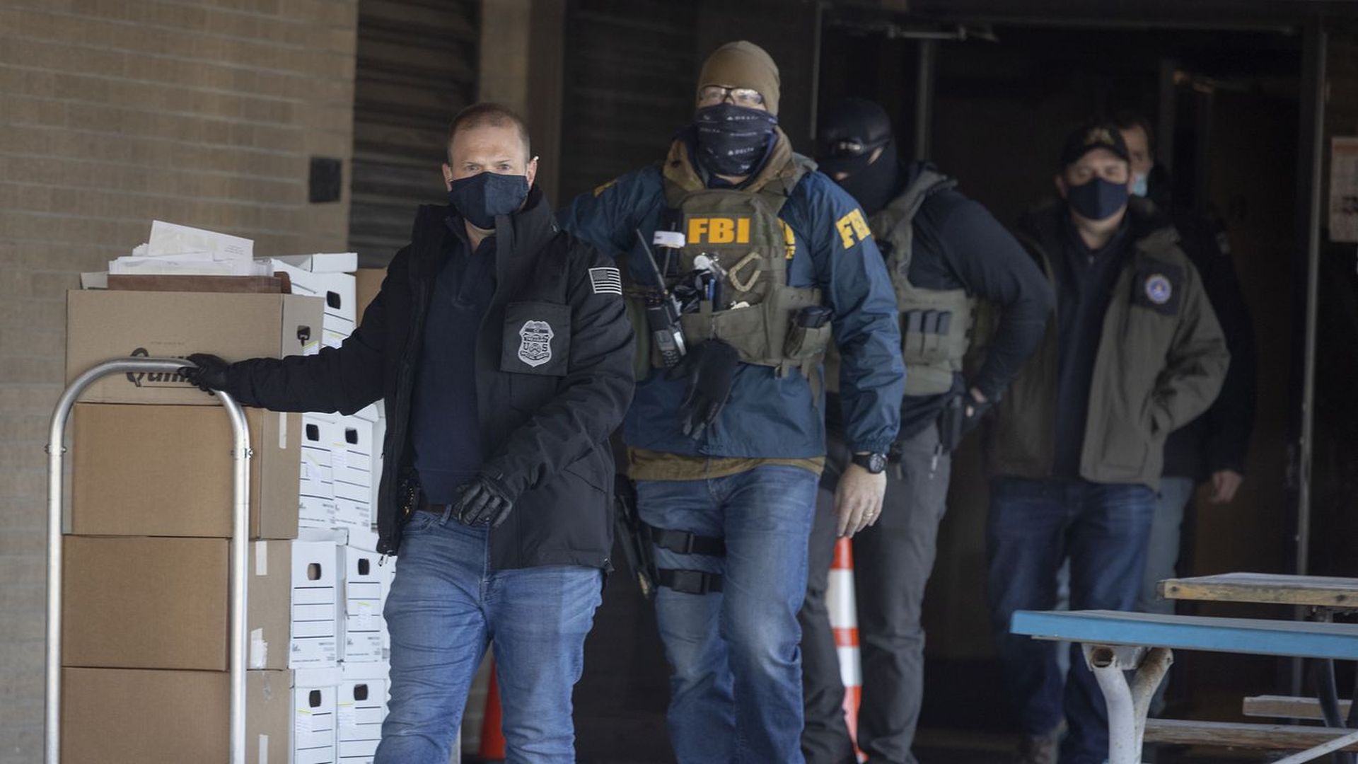 Law enforcement agents in blue jeans and masks walk with a cart stacked high with boxes
