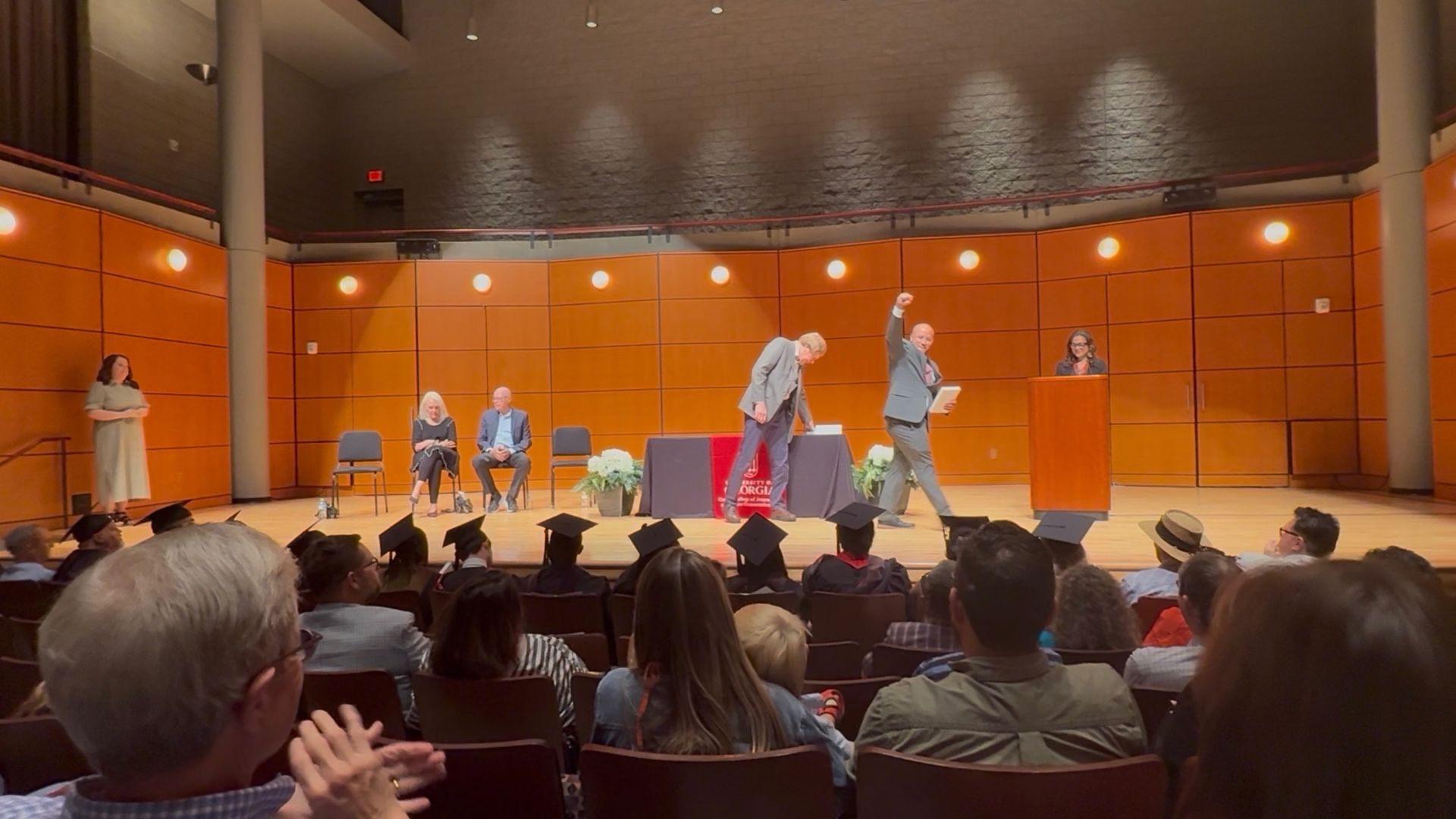 Stage with orange paneled walls; two men in suits center, one raising a fist. A woman speaks at the podium right; seated panelists and an audience in graduation caps fill the foreground.