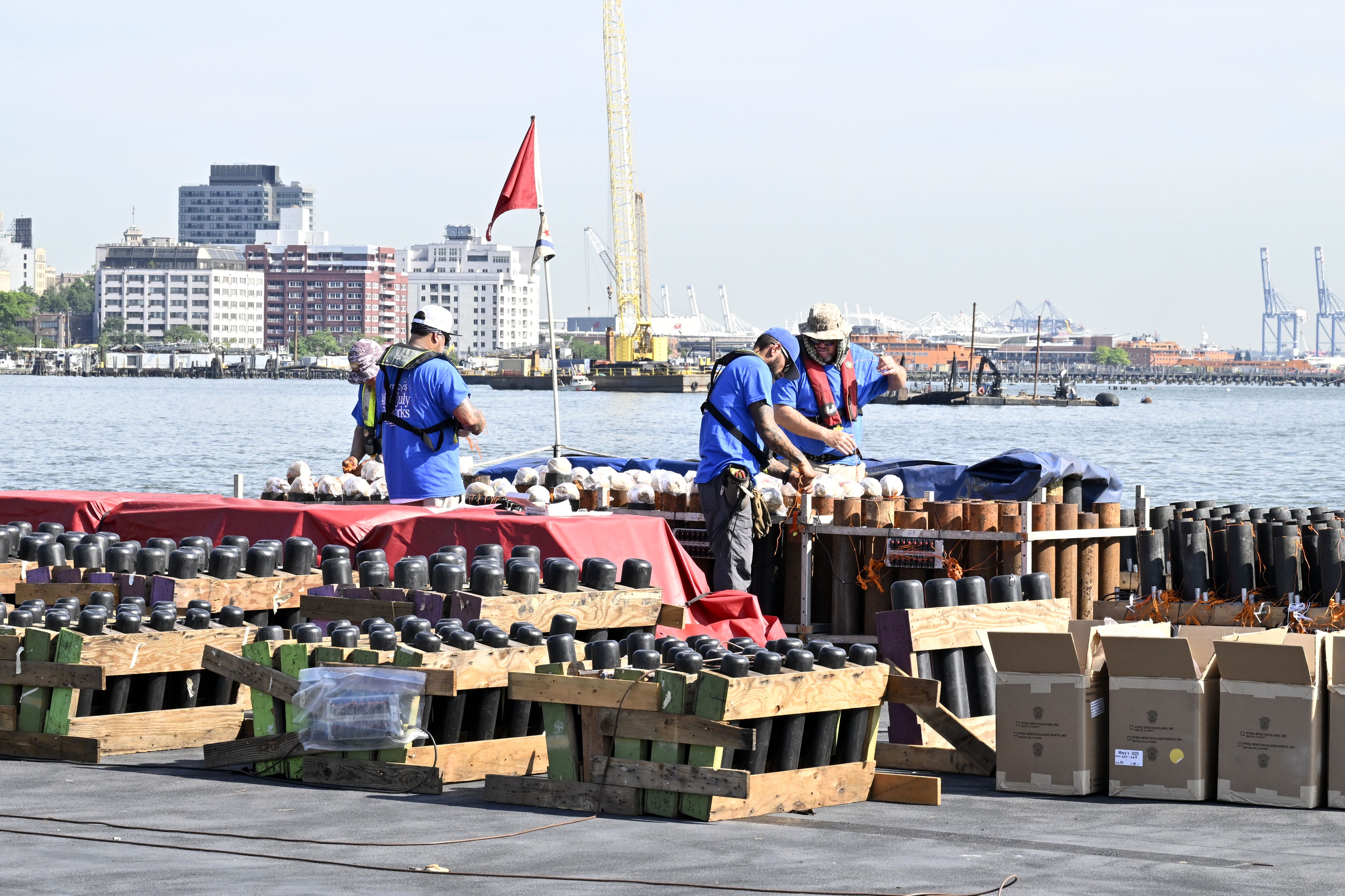 Three workers in blue shirts preparing fireworks on a dock by the water with a city skyline and cranes in the background during daytime.