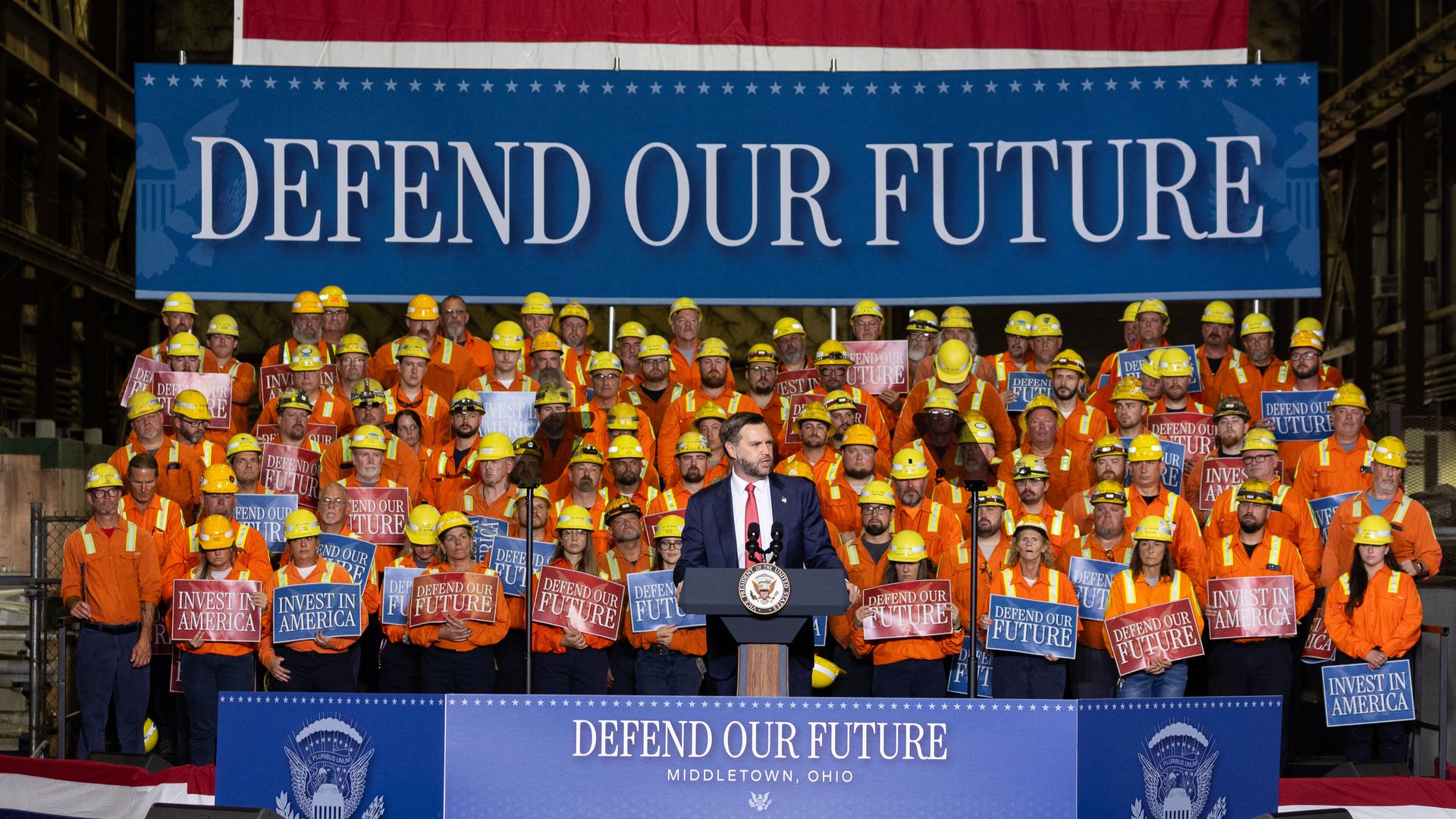 US Vice President JD Vance stands at a podium while delivering remarks on the Trump Administration's "America First" economic agenda at the Cleveland-Cliffs Middletown Works steel mill in Middletown, Ohio. A couple dozen steelworkers on rafters hold signs behind him.
