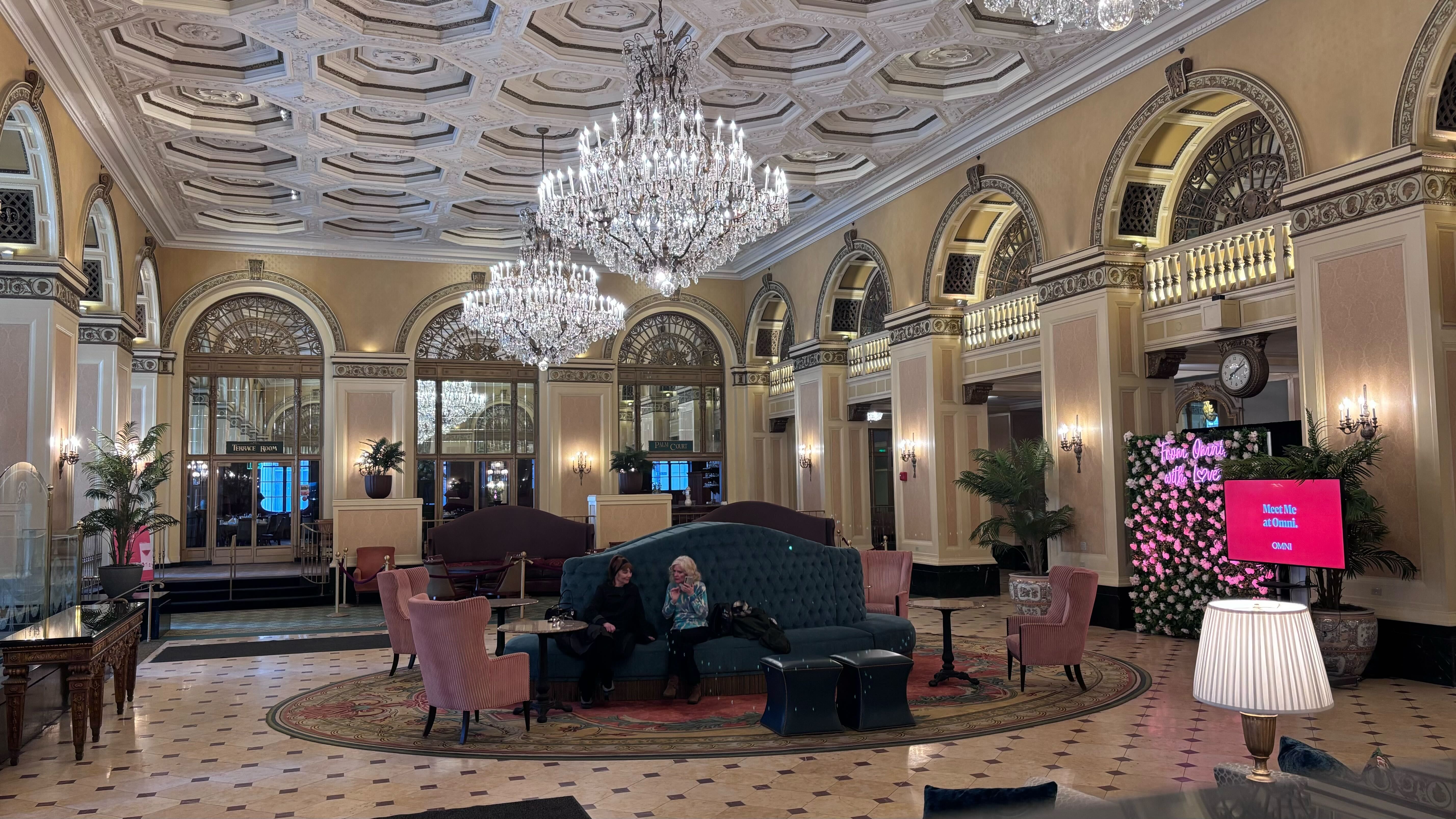 Luxurious hotel lobby with ornate white ceiling, large crystal chandeliers, beige walls with arched windows, velvet seating, two women conversing, and a pink floral wall with a neon sign.
