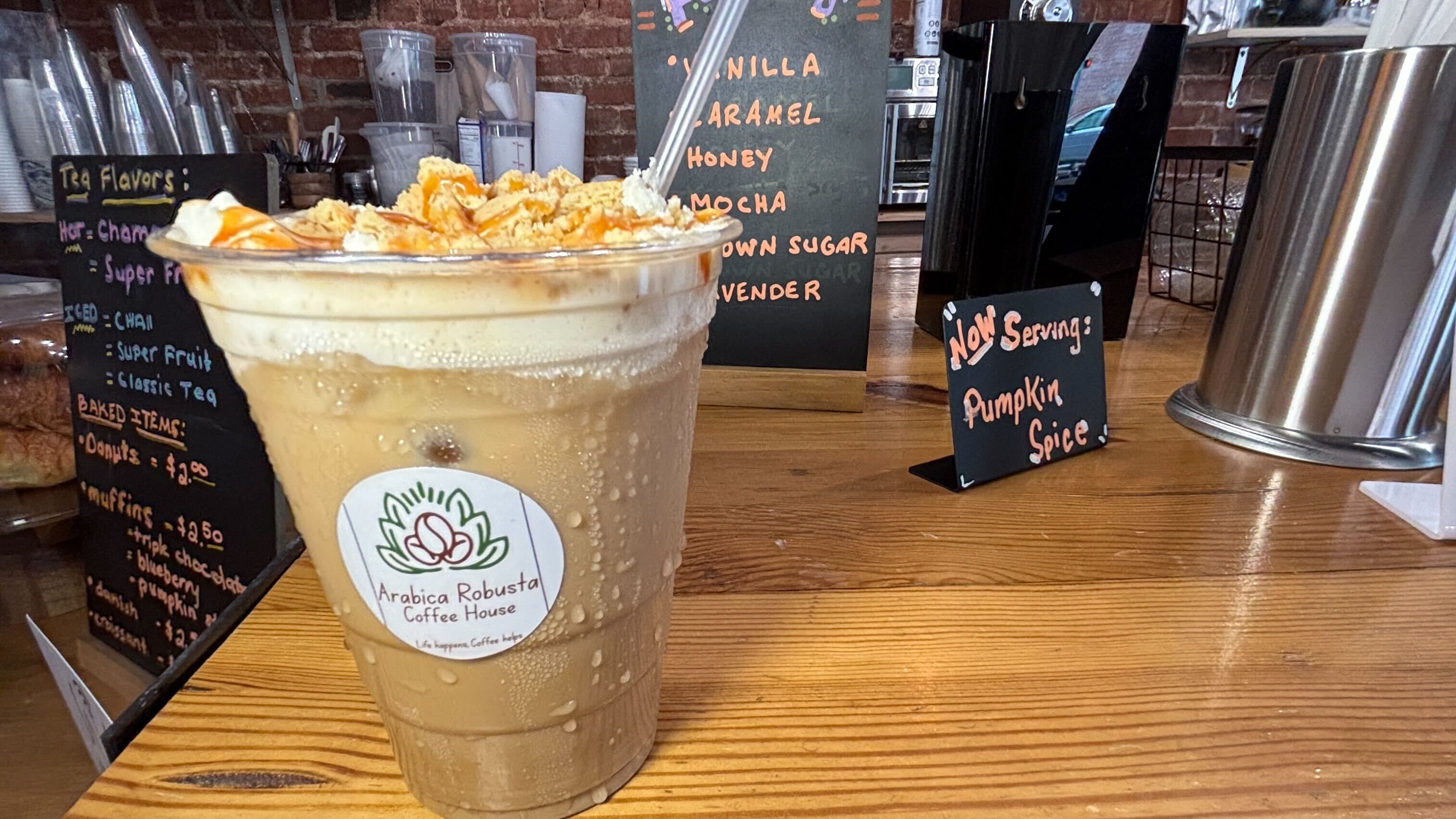 Iced coffee drink with whipped cream and caramel drizzle in plastic cup labeled Arabica Robusta Coffee House, on wooden counter with signs showing tea flavors and pumpkin spice availability.