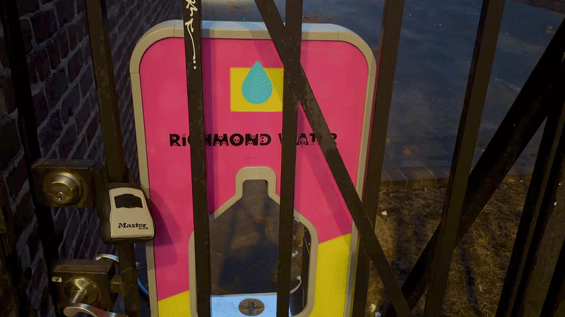 a video of a hand with a water bottle filling up at a refill station behind a fence.