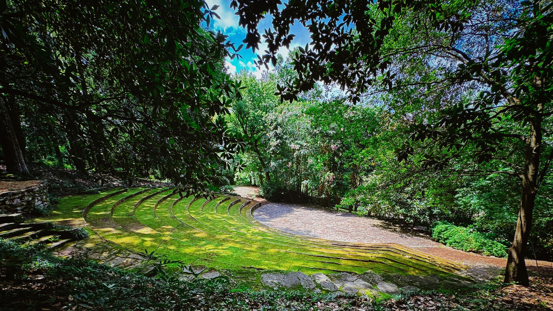 Outdoor amphitheater with curved stone steps covered in green moss, surrounded by lush trees and foliage under a partly cloudy blue sky.