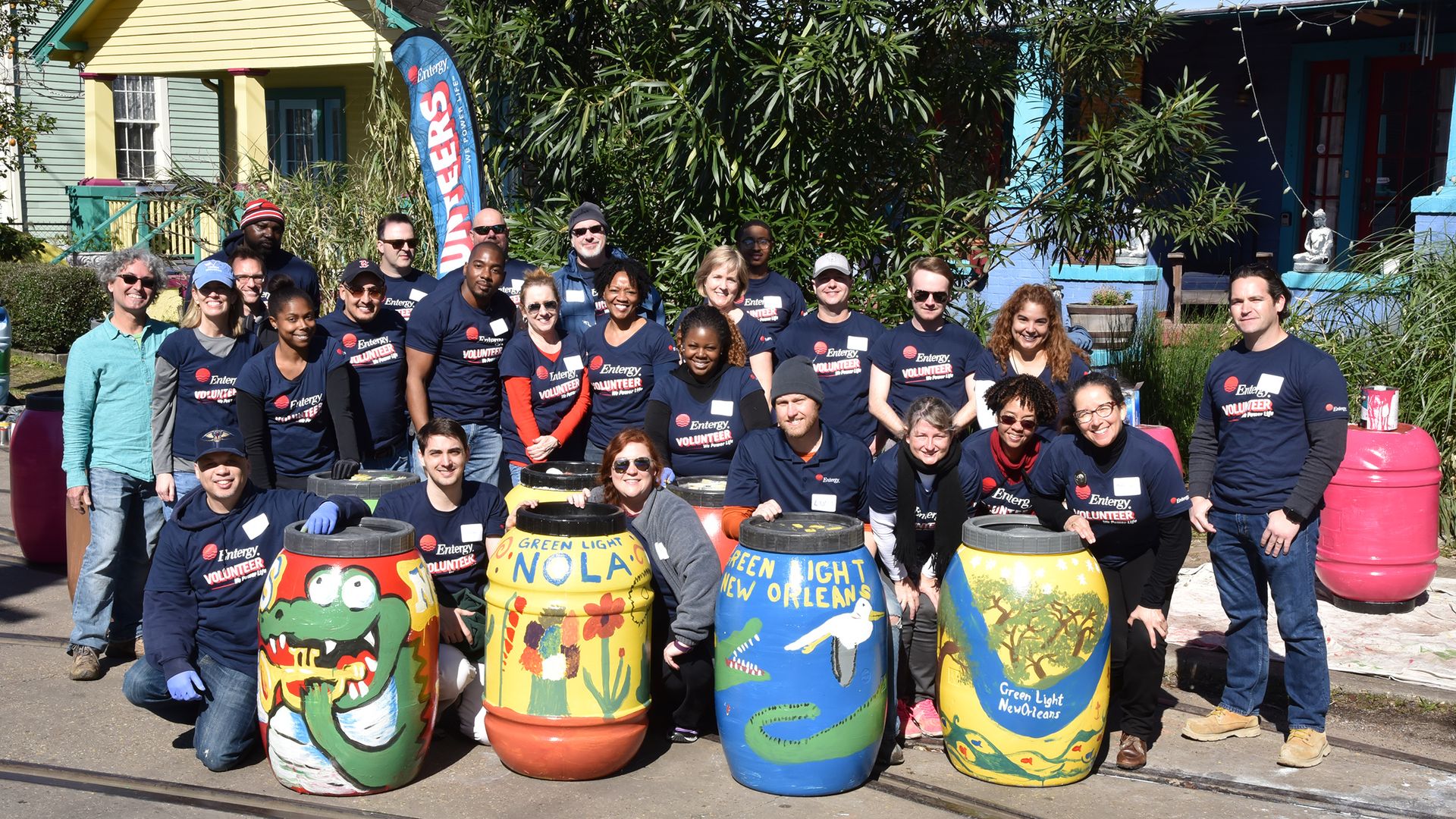 Group of volunteers wearing navy shirts posing outdoors with colorful painted barrels labeled "Green Light New Orleans" in front of houses and greenery on a sunny day.