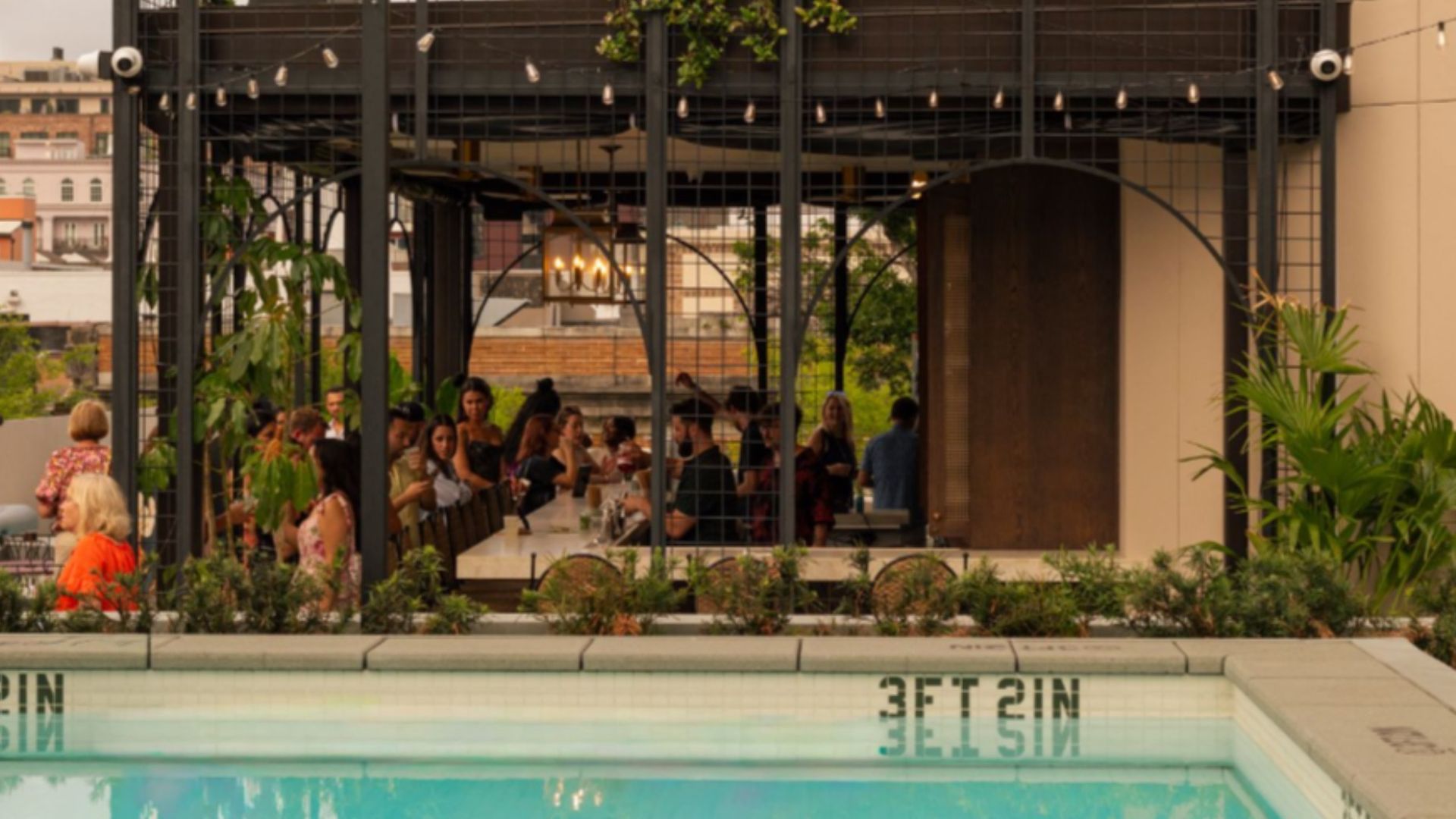 Outdoor rooftop dining under a black metal pergola with string lights, a long white table full of people, plants around, warm lighting; city buildings in the background and a pool in the foreground.