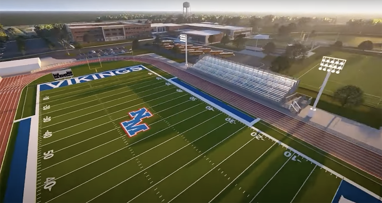 Aerial view of a green American football field with "VIKINGS" in white on a blue background in one end zone and a large blue and red "M" logo at midfield. Bleachers and stadium lights visible.