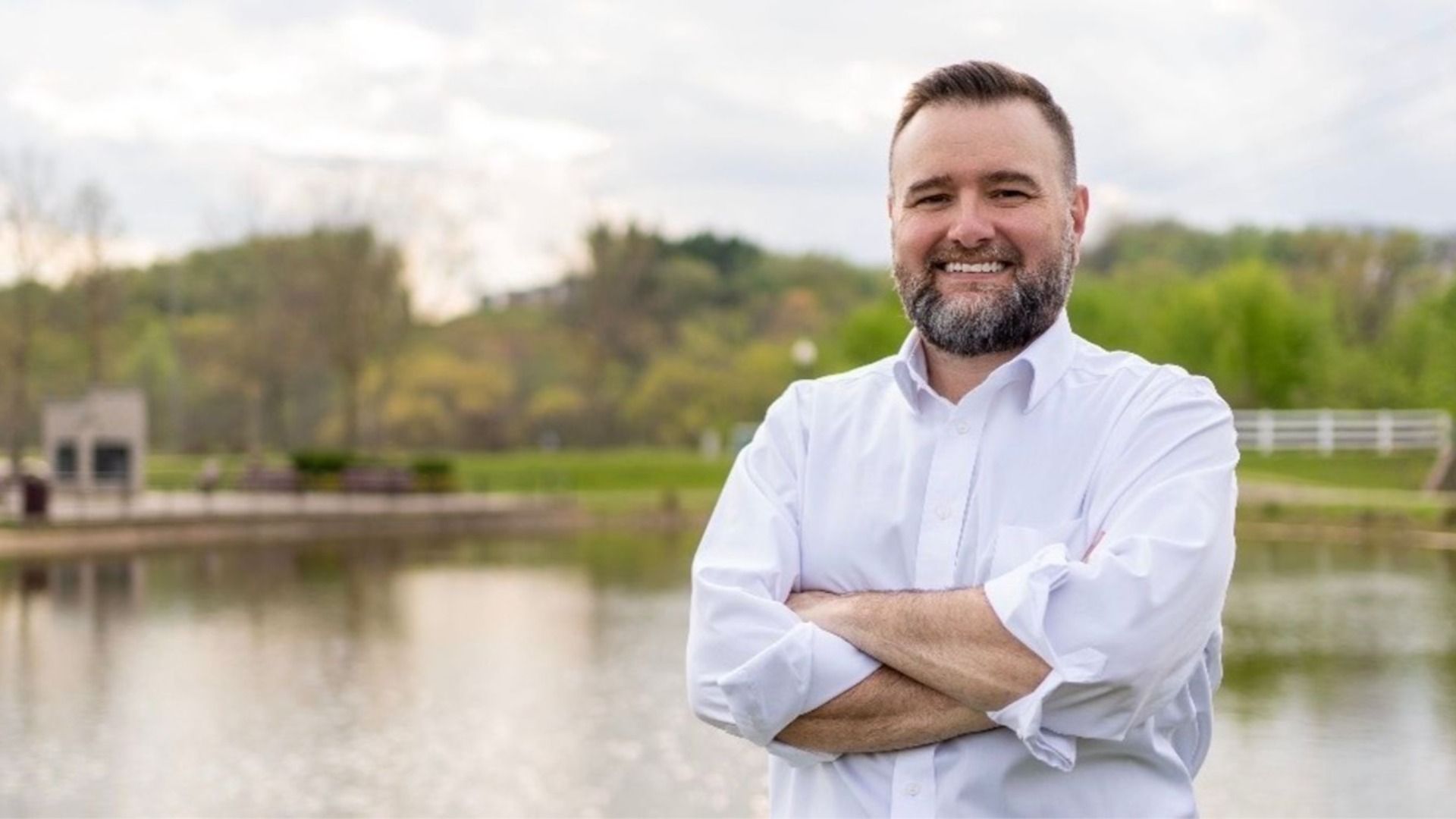 Smiling man with a beard wearing a white button-down shirt stands with arms crossed beside a lake in a park; blurred trees and a fence in the background.
