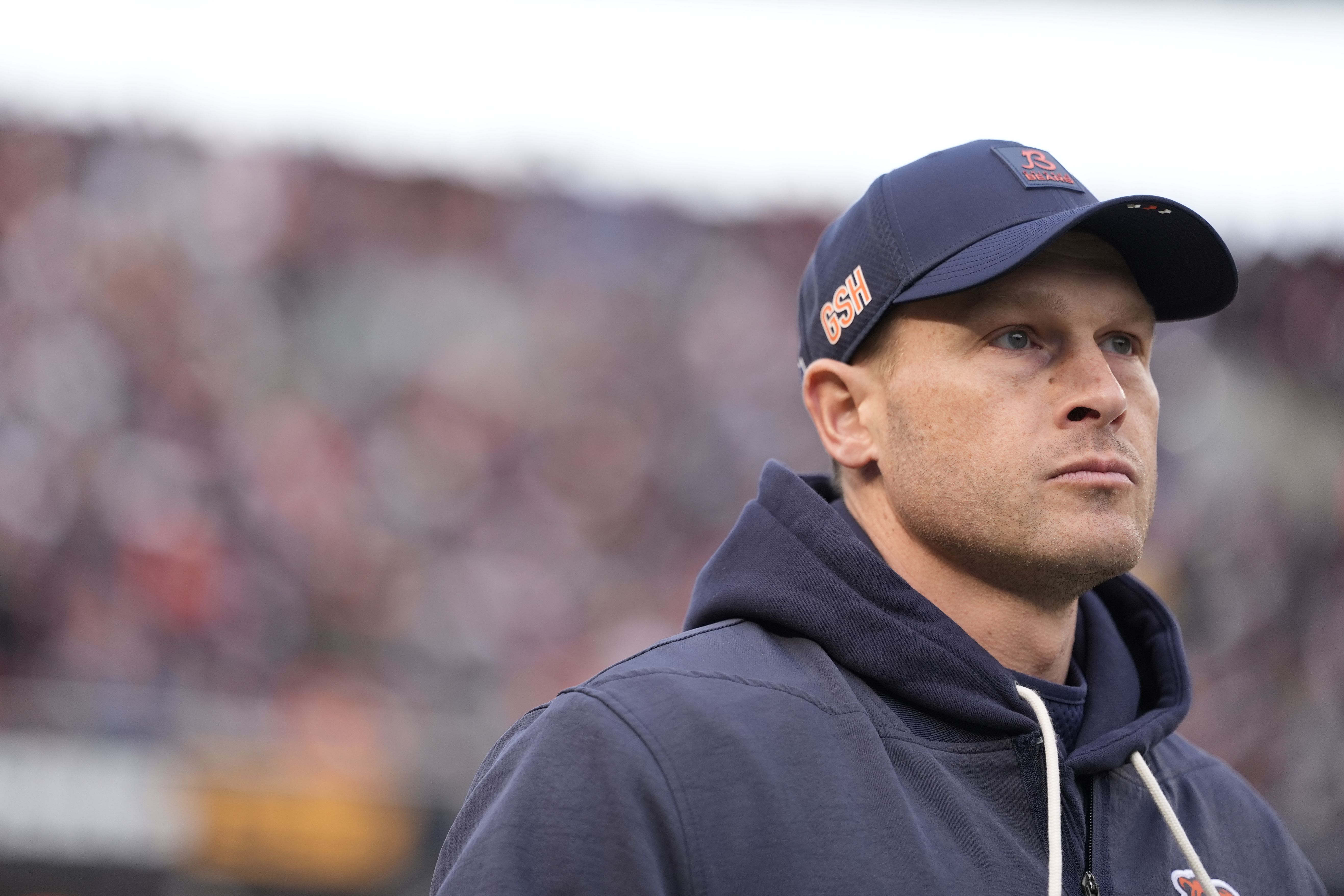Man wearing a navy blue cap with orange lettering and a navy blue hoodie looks ahead with a serious expression, blurred crowd in the background.