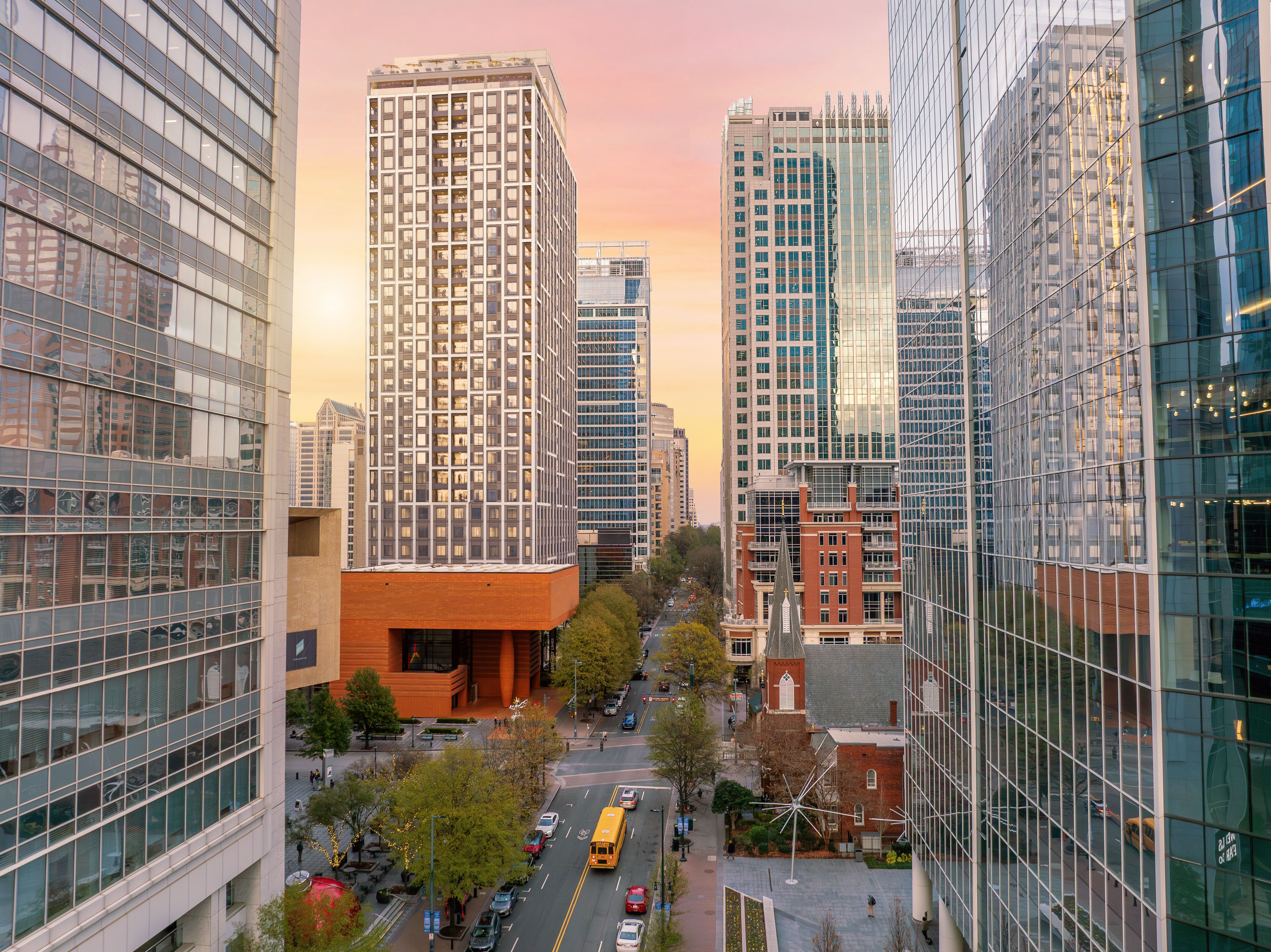 City street canyon between glass skyscrapers at sunset; warm orange-pink sky, yellow school bus and cars on the road, trees lining sidewalks.