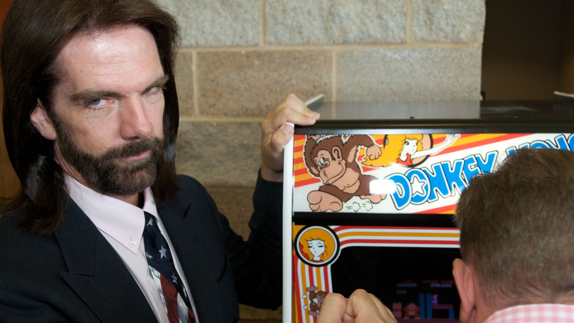 Billy Mitchell, with long dark hair and beard, stands beside a Donkey Kong arcade game. 