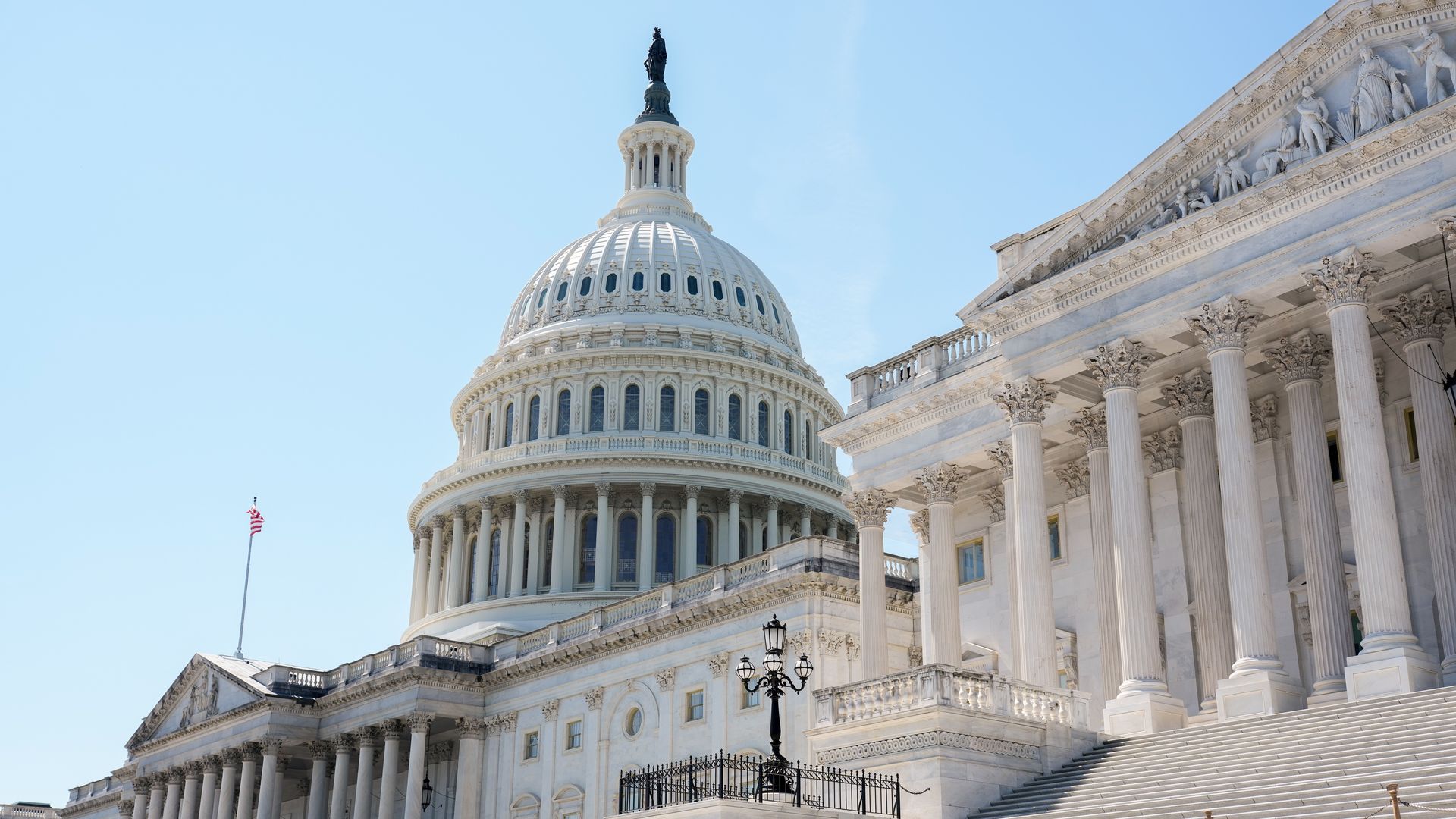 White neoclassical capitol building with a large domed roof topped by a statue, flanked by columned facades and long marble steps, set against a clear blue sky.