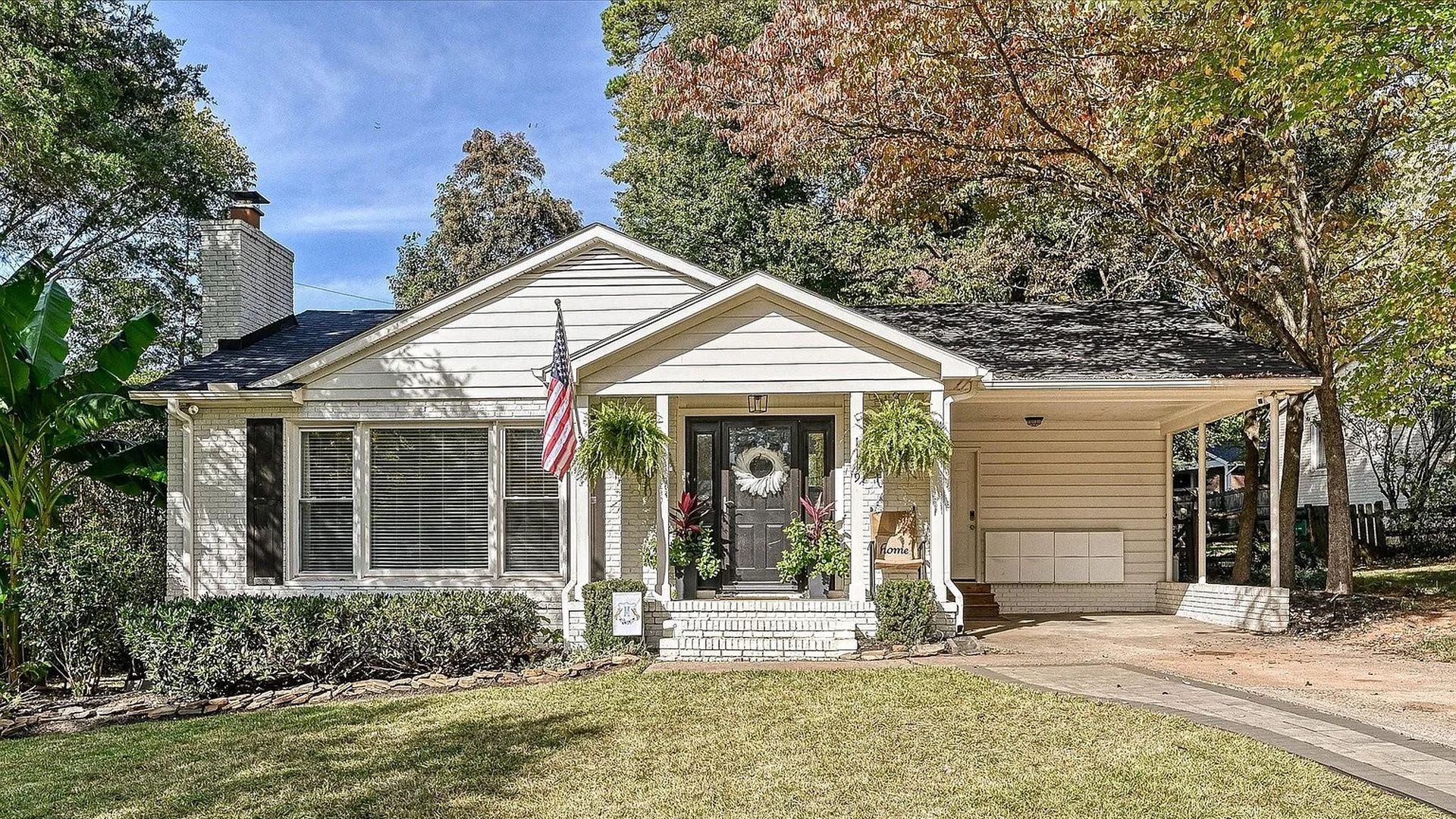 White single-story house with black front door, American flag, two hanging ferns, green shrubs, and a large front lawn under a partly cloudy blue sky in autumn.