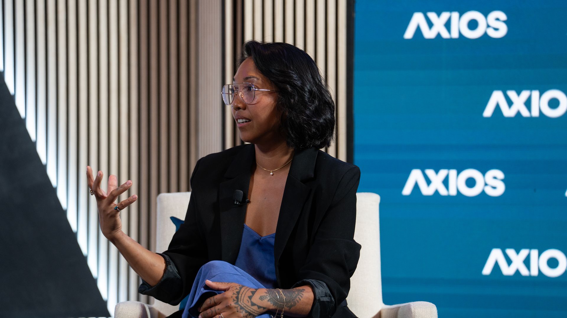 Side-profile of a woman with round glasses, wearing a black blazer and blue dress, gesturing with her tattooed forearm while seated on a beige chair; a blue Axios backdrop behind her.