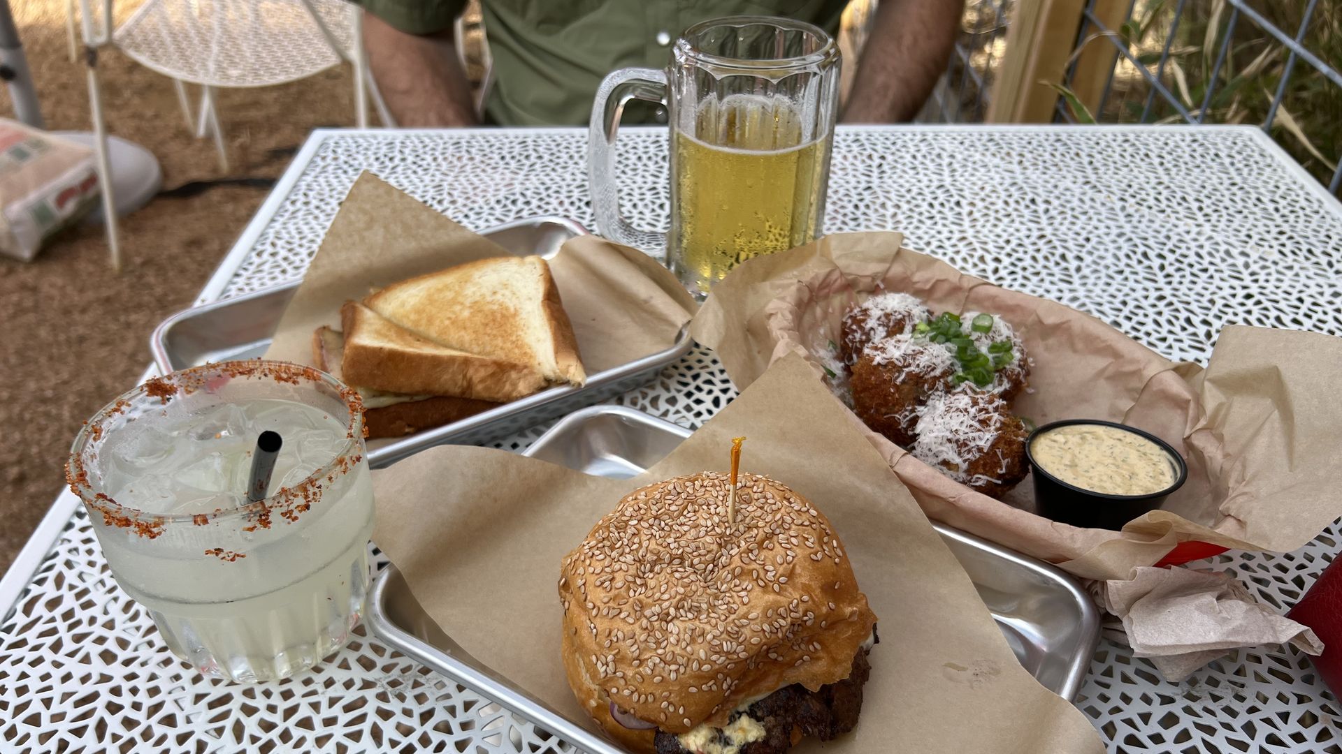 Outdoor table with a sesame-seed hamburger on parchment, a tray of toasted sandwich triangles, a frosty beer mug, and two cheese-topped fried croquettes with dipping sauce, on a white lattice table.