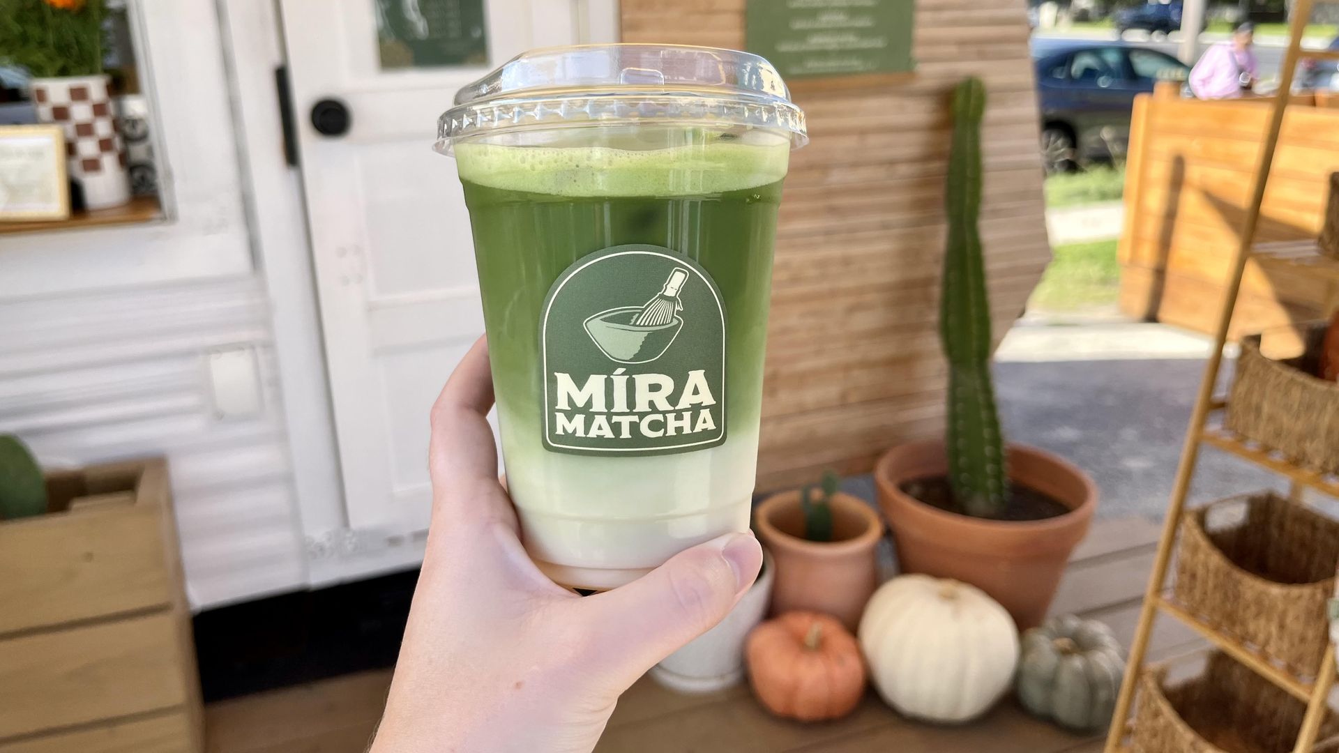 A hand holding a clear plastic cup of green matcha layered over white milk. The cup is labeled "MÍRA MATCHA" with a green sticker. Blurred behind it is a cozy fall setting with pumpkins and cacti.