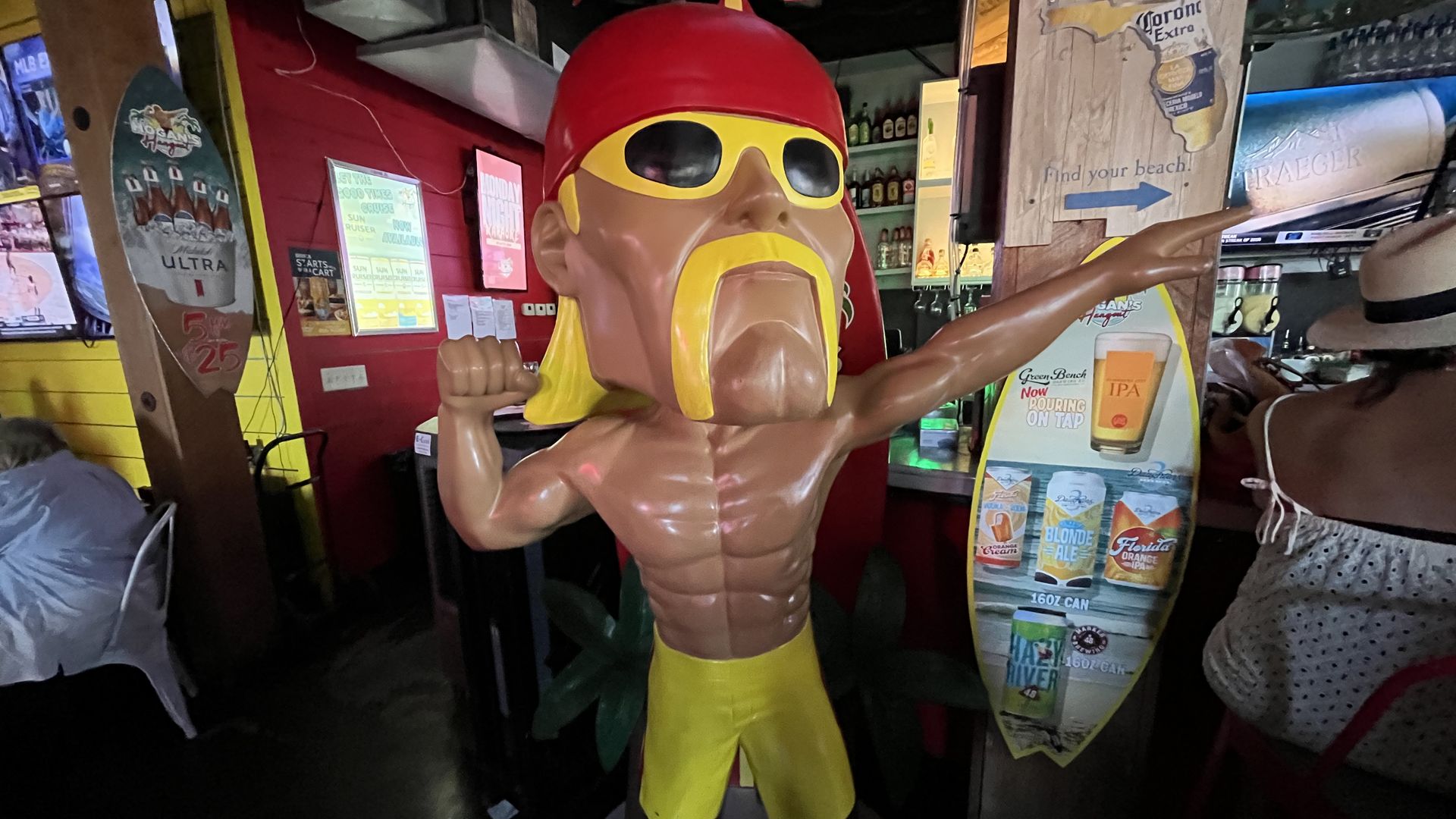Statue of a muscular man with yellow sunglasses, yellow mustache and beard, red cap, and yellow shorts and flip-flops, posing indoors near a surfboard and bar counter.