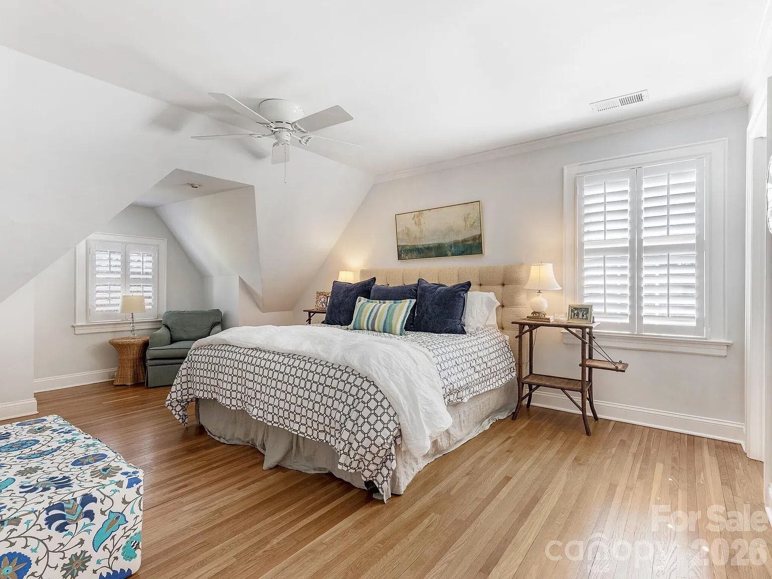 Bright bedroom with hardwood floor, white walls, large bed with beige tufted headboard, patterned bedspread, blue and striped pillows, ceiling fan, side tables with lamps, and shuttered windows.