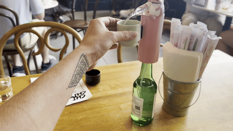 A hand reaches a small white cup toward a drink dispenser attached to the top of a soju bottle.