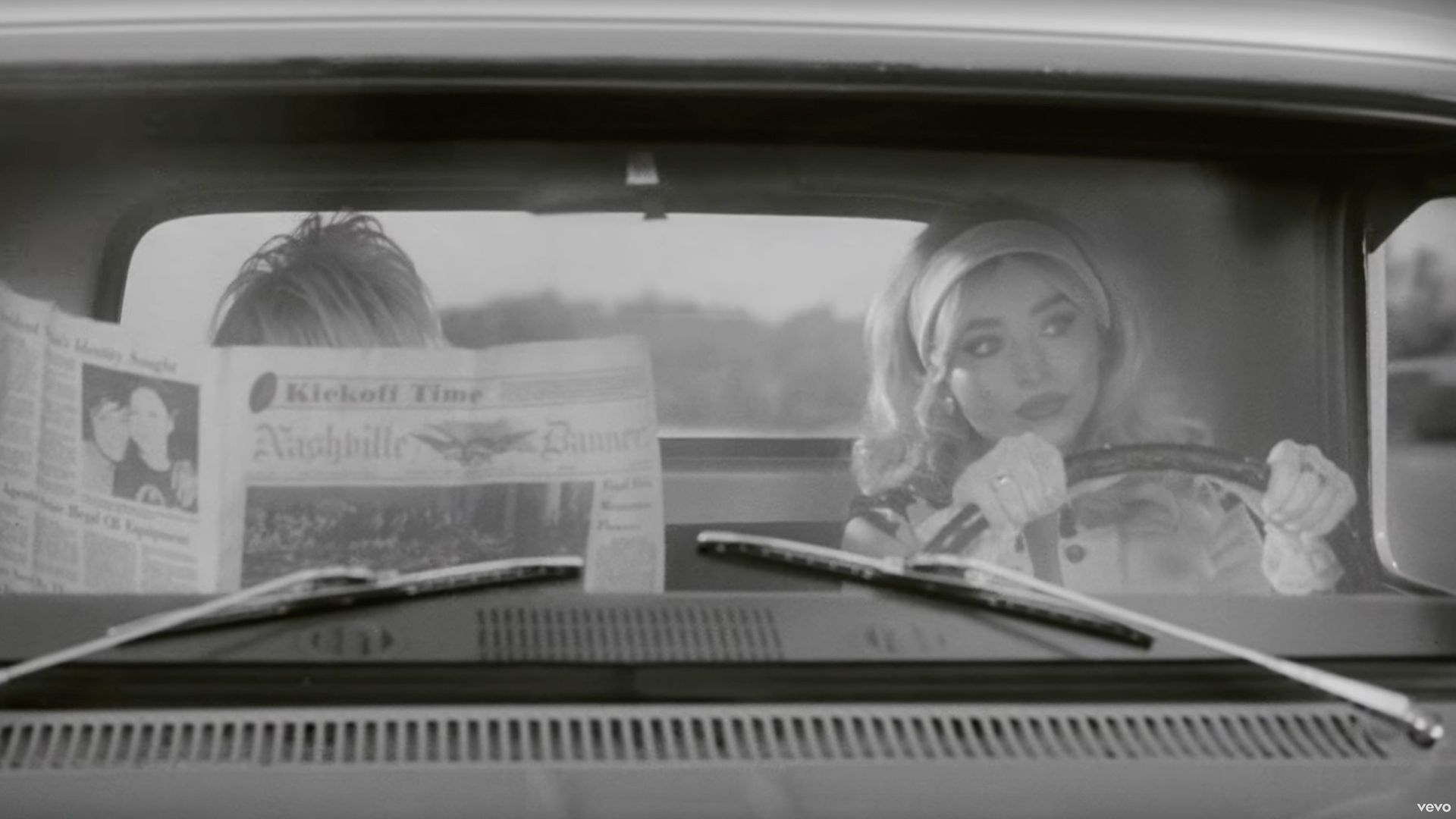 Dolly Parton reading a newspaper in a truck while Sabrina Carpenter drives.