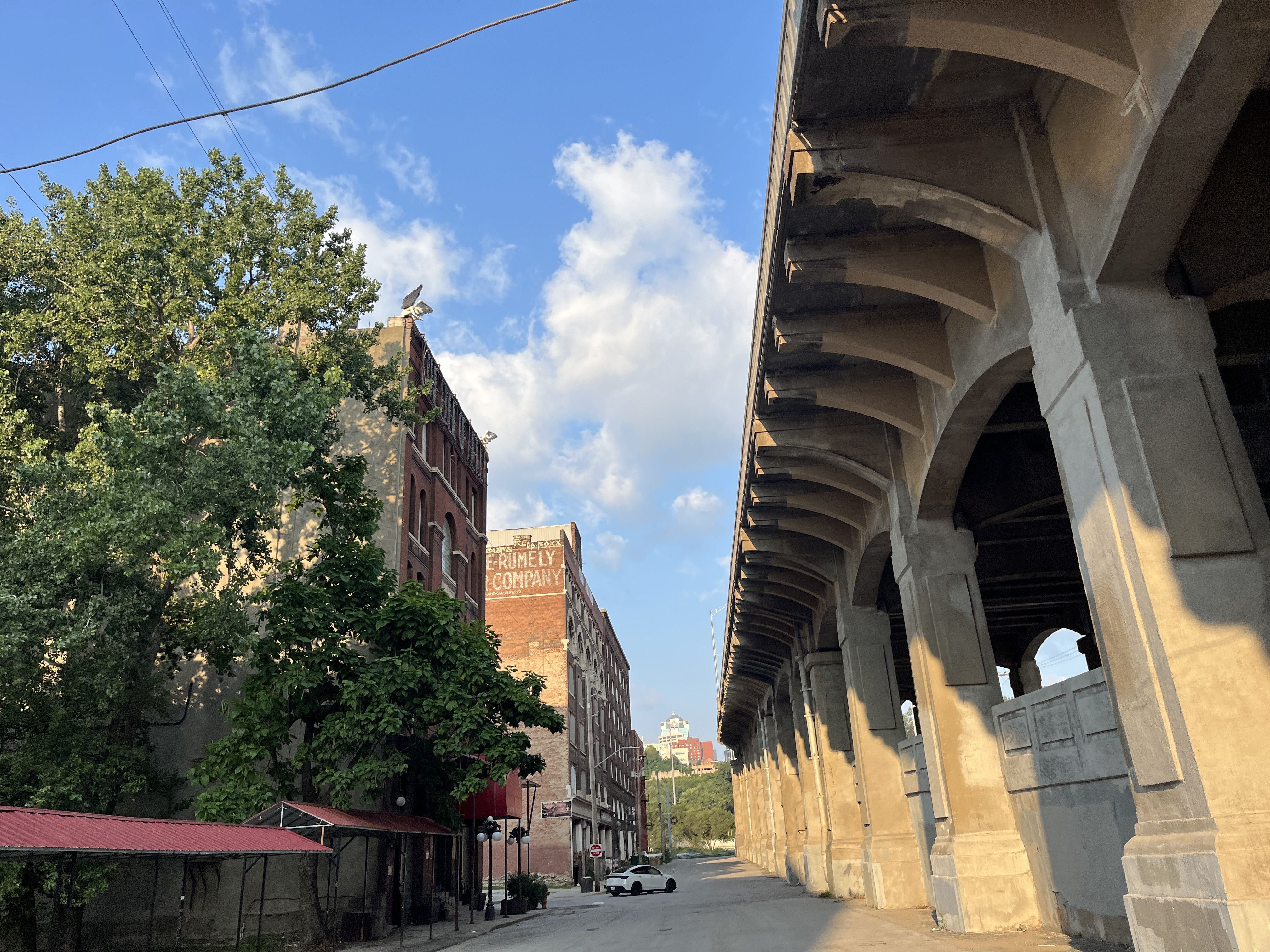 Street scene with brick buildings, one with faded "Rumely Company" sign, tall green trees, and a large concrete elevated structure casting shadows under a partly cloudy blue sky.