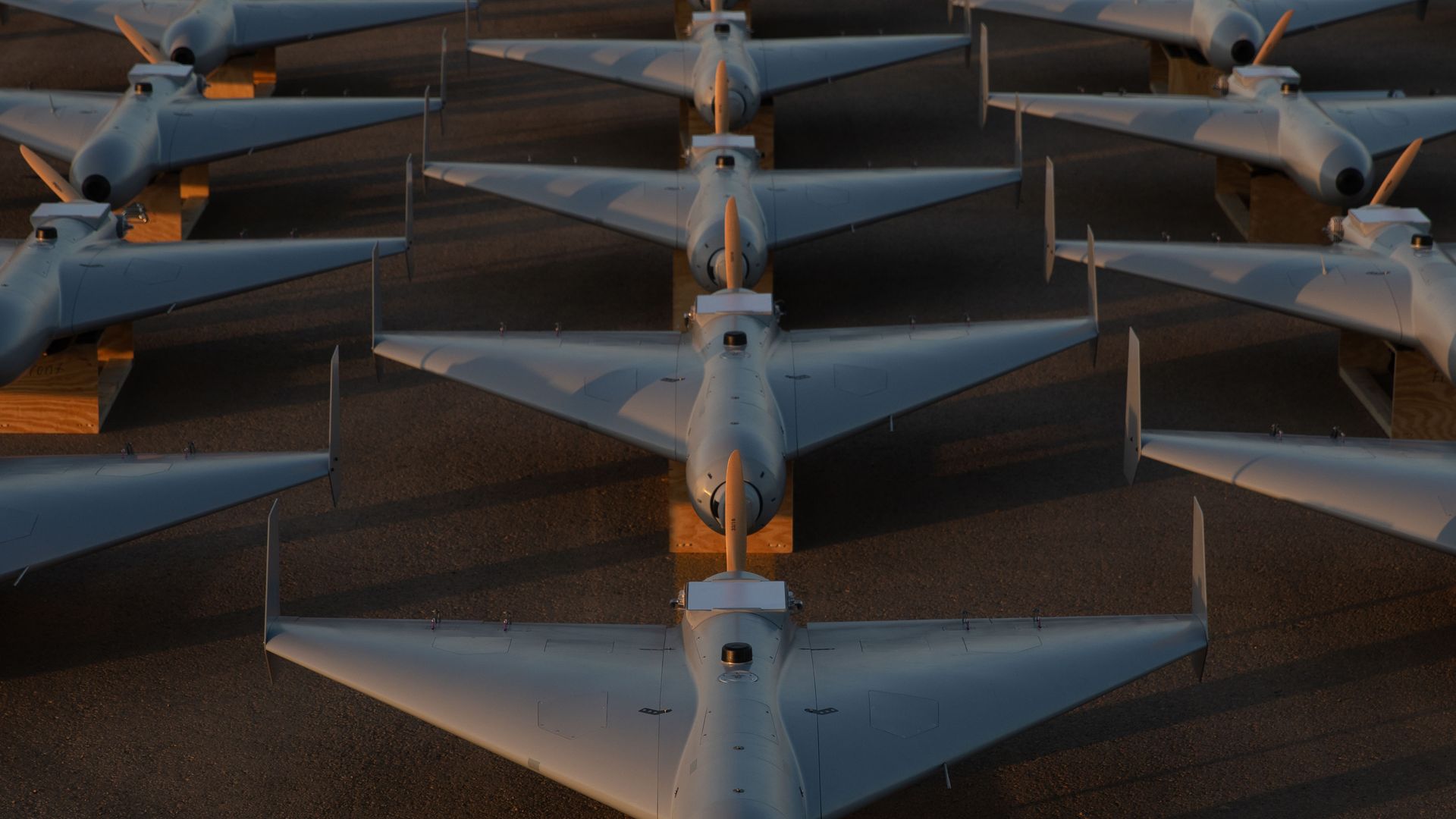 Rows of gray drones arranged in a grid on a paved surface, lit by soft sunlight casting shadows between them.