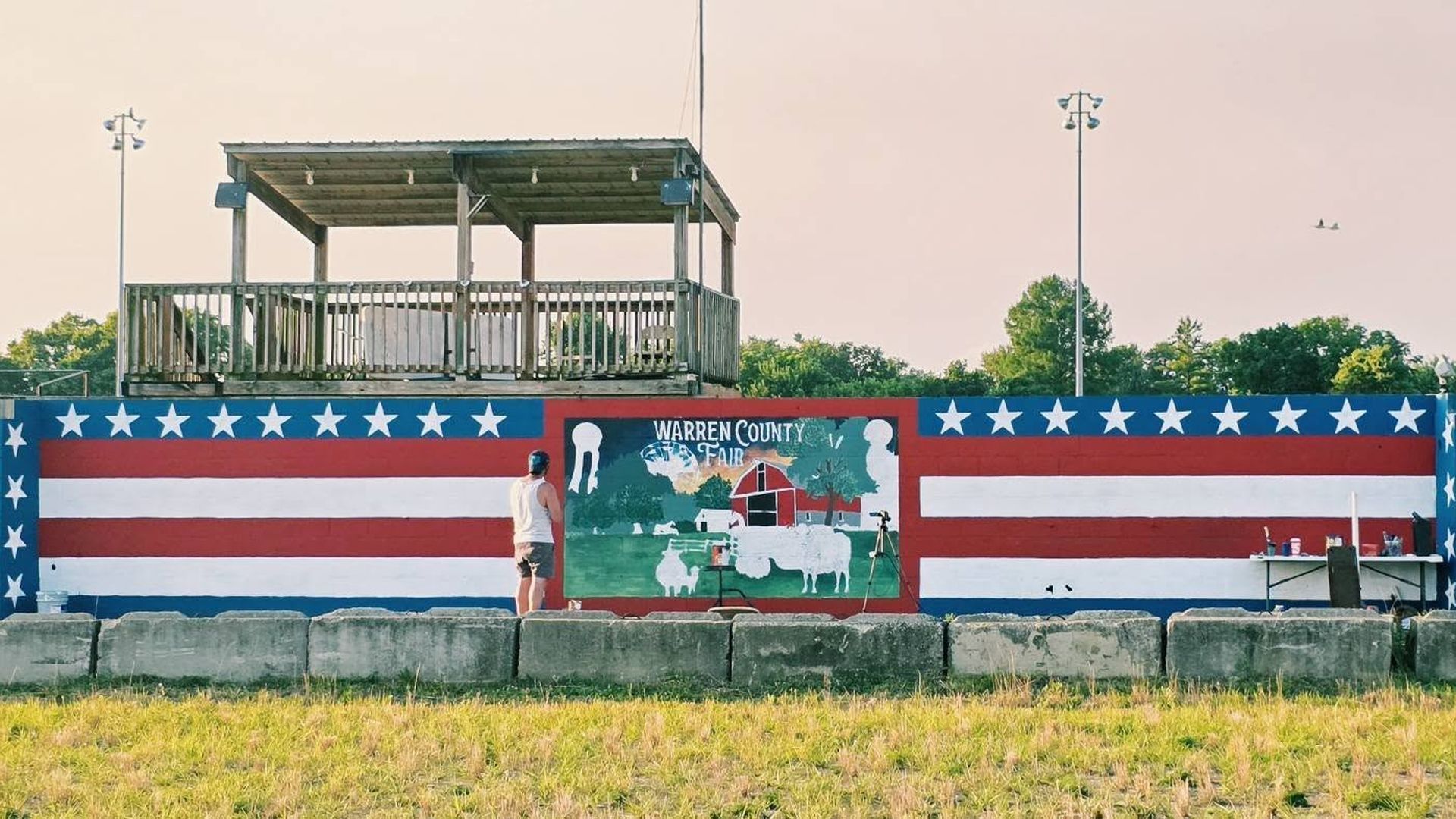 Patriotic mural with red, white, and blue stripes and white stars; center panel reads "Warren County Fair" and shows a farm scene with a red barn. A man in a white tank top paints in front, beneath a wooden viewing deck.