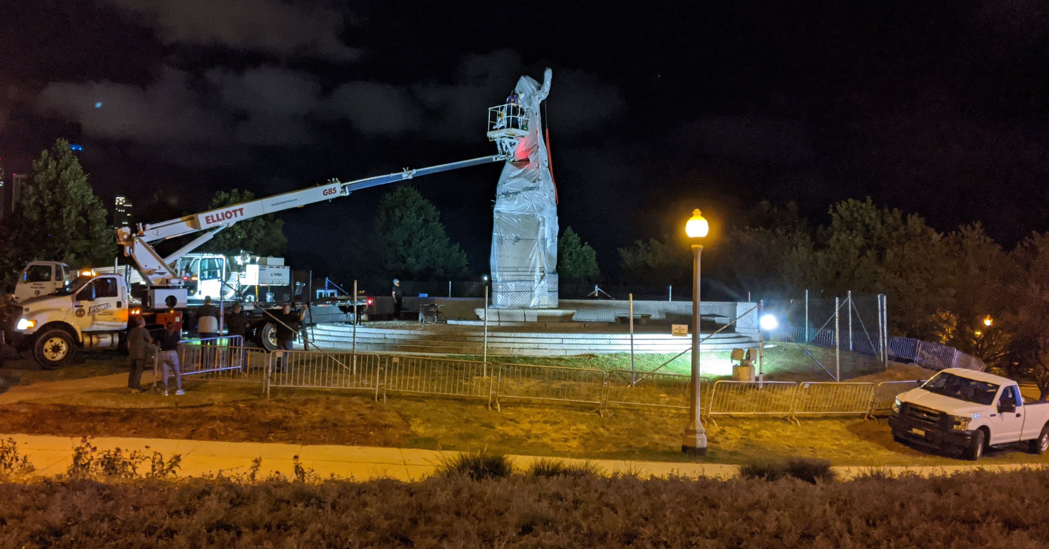 A crane removes a large Columbus monument in the dark. The pedestal is fenced in.