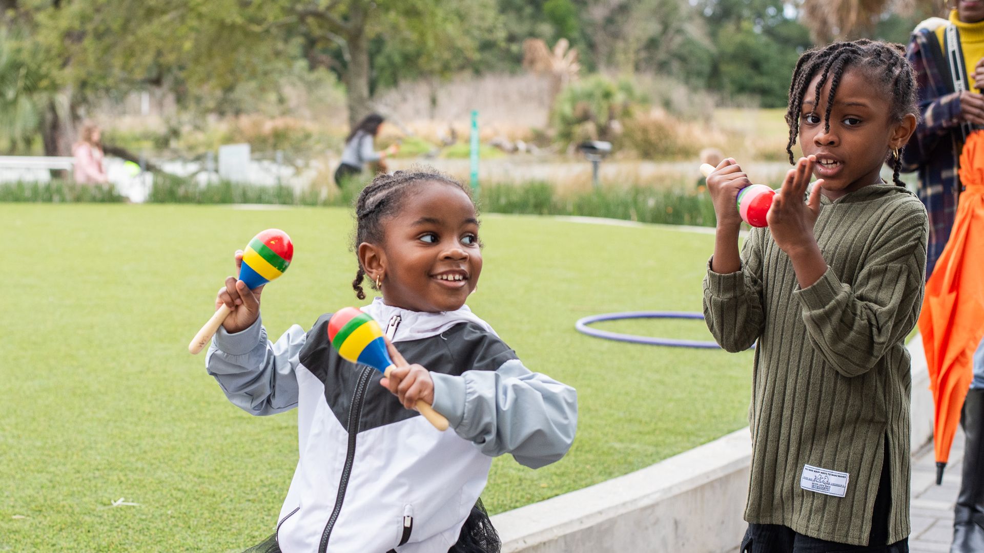 Two young girls play with colorful maracas outdoors in a park. One wears a gray and white jacket, the other a green ribbed top. A person in yellow stands in the background near trees and grass.
