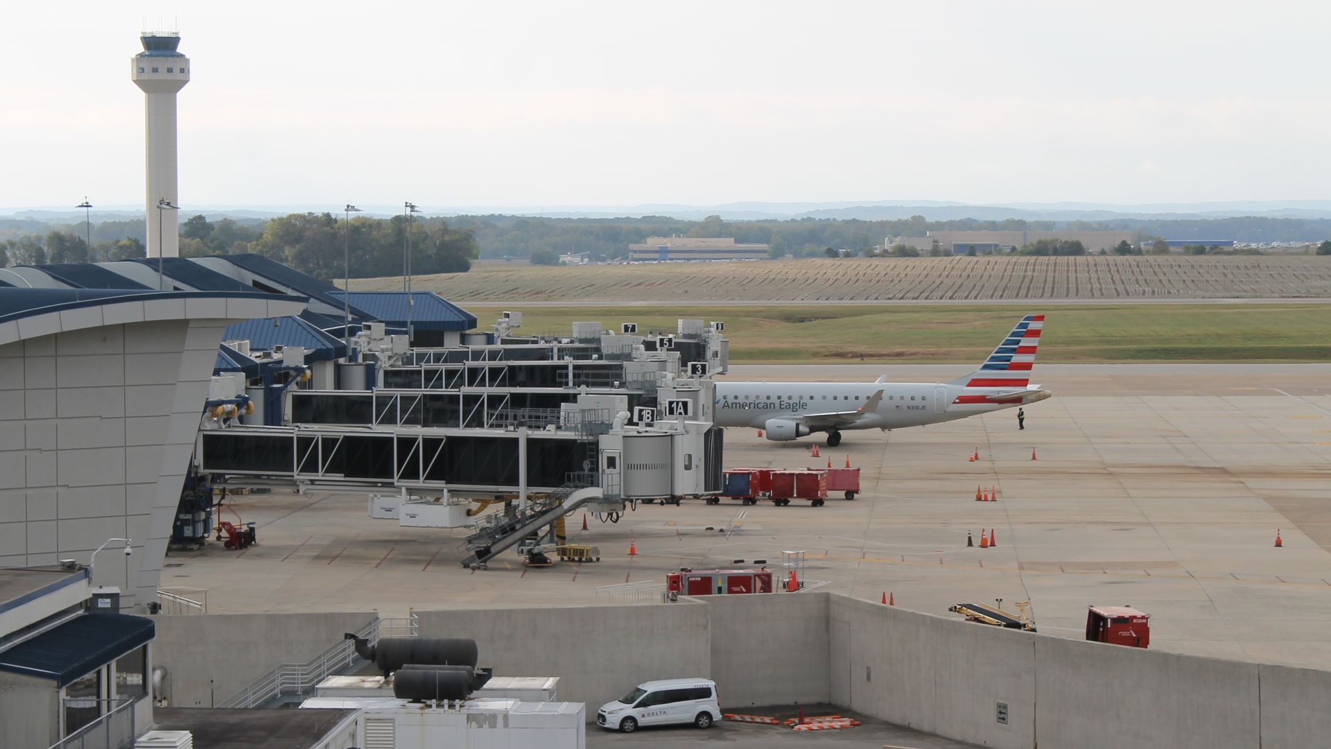 Airport terminal with boarding gates, jet bridges, an American Eagle airplane, a control tower, parked service vehicles, and a runway extending to fields under a cloudy sky.