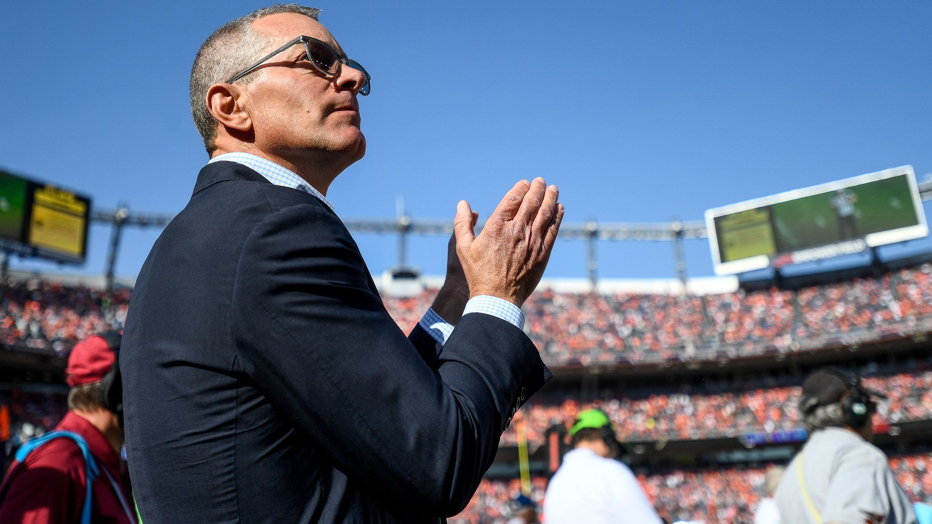 Side-profile of a mature man in a dark suit and sunglasses, clapping at a sunny stadium with a sea of orange-clad fans and large video boards in the background.