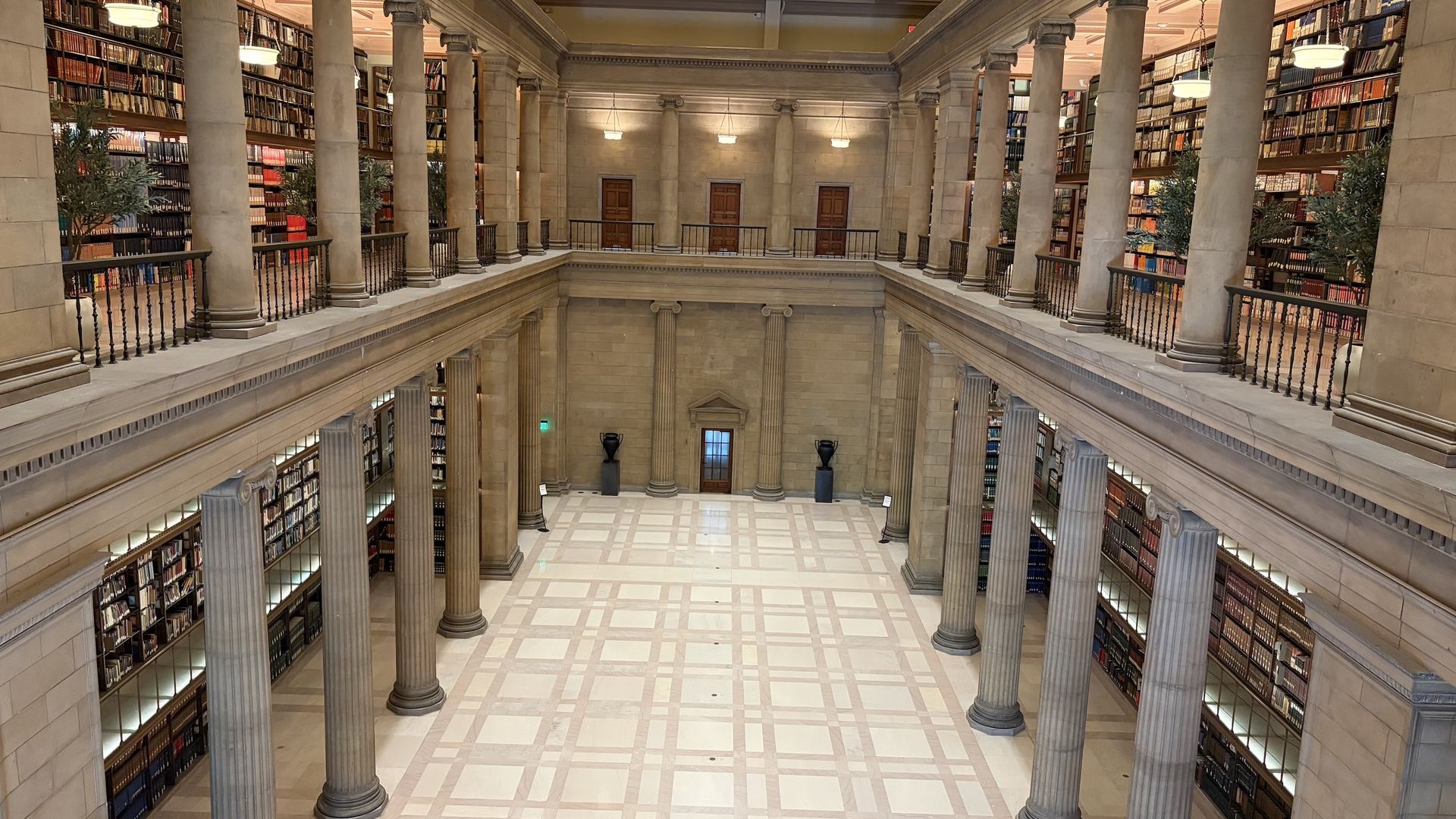 Interior of a grand library with two levels of bookshelves, tall stone columns, iron railings, and warm lighting. A skylit ceiling and beige marble floors create a classical, airy atrium.
