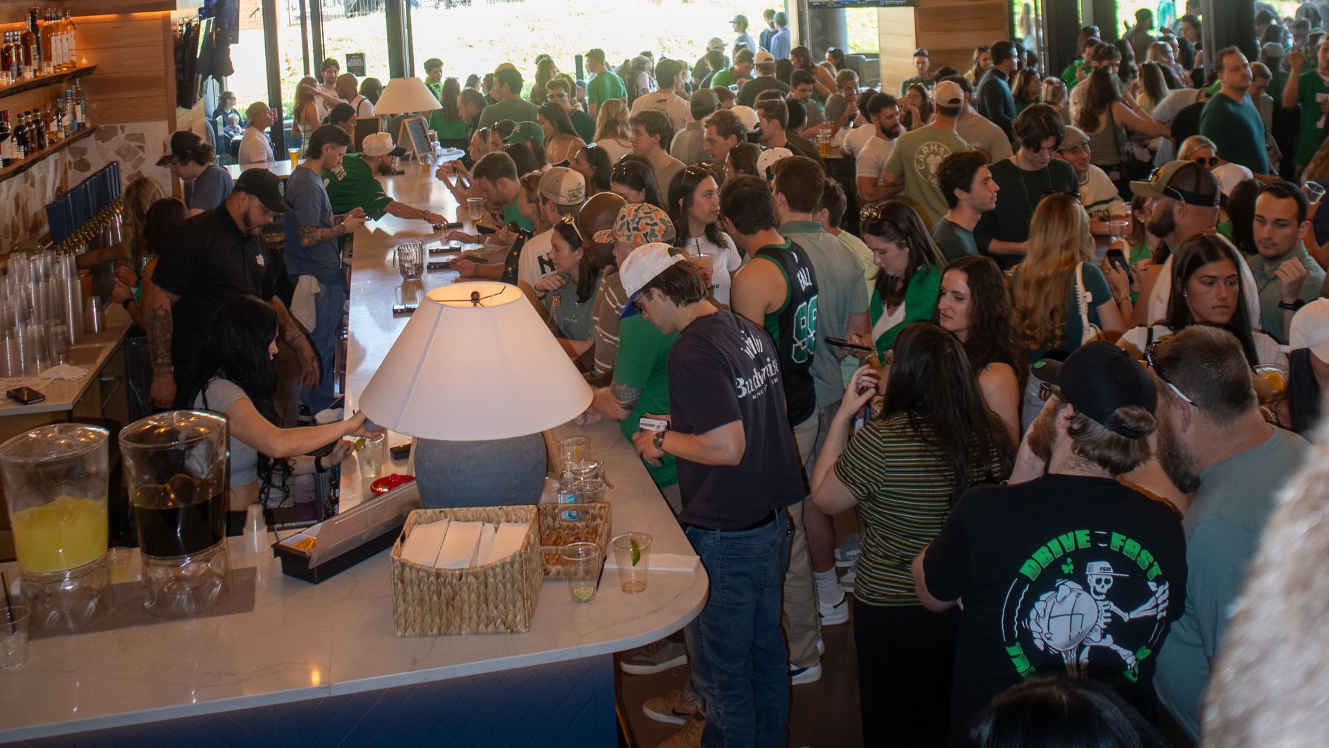 Busy cafe interior with a long counter crowded with patrons. Large circular chandeliers cast warm light over an exposed-duct ceiling, while groups mingle near windows in the back.