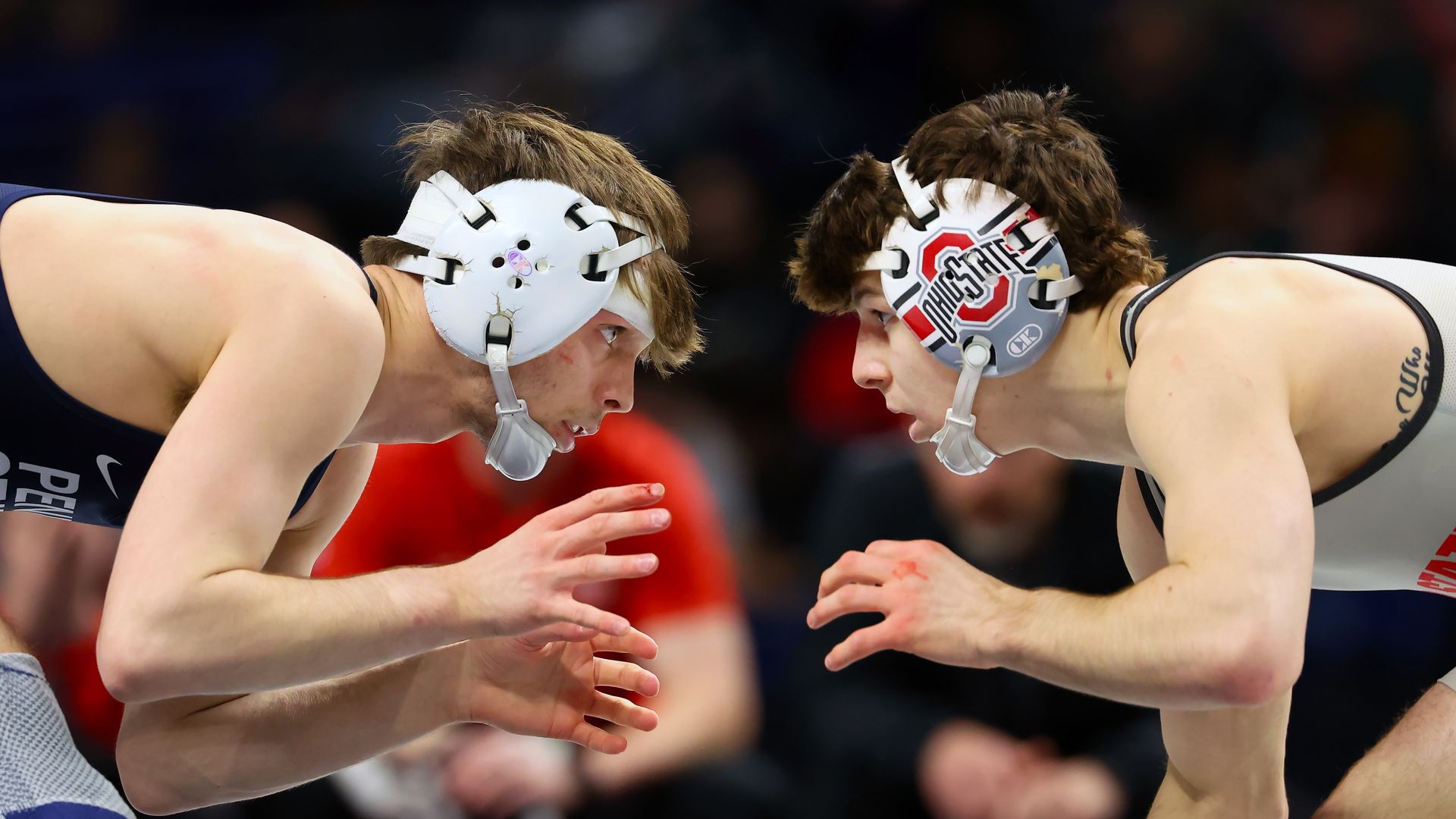 Two male wrestlers crouch in a close standoff on the mat, wearing white headgear. Left in a dark blue singlet, right in a gray singlet with an Ohio State emblem; hands raised and ready.