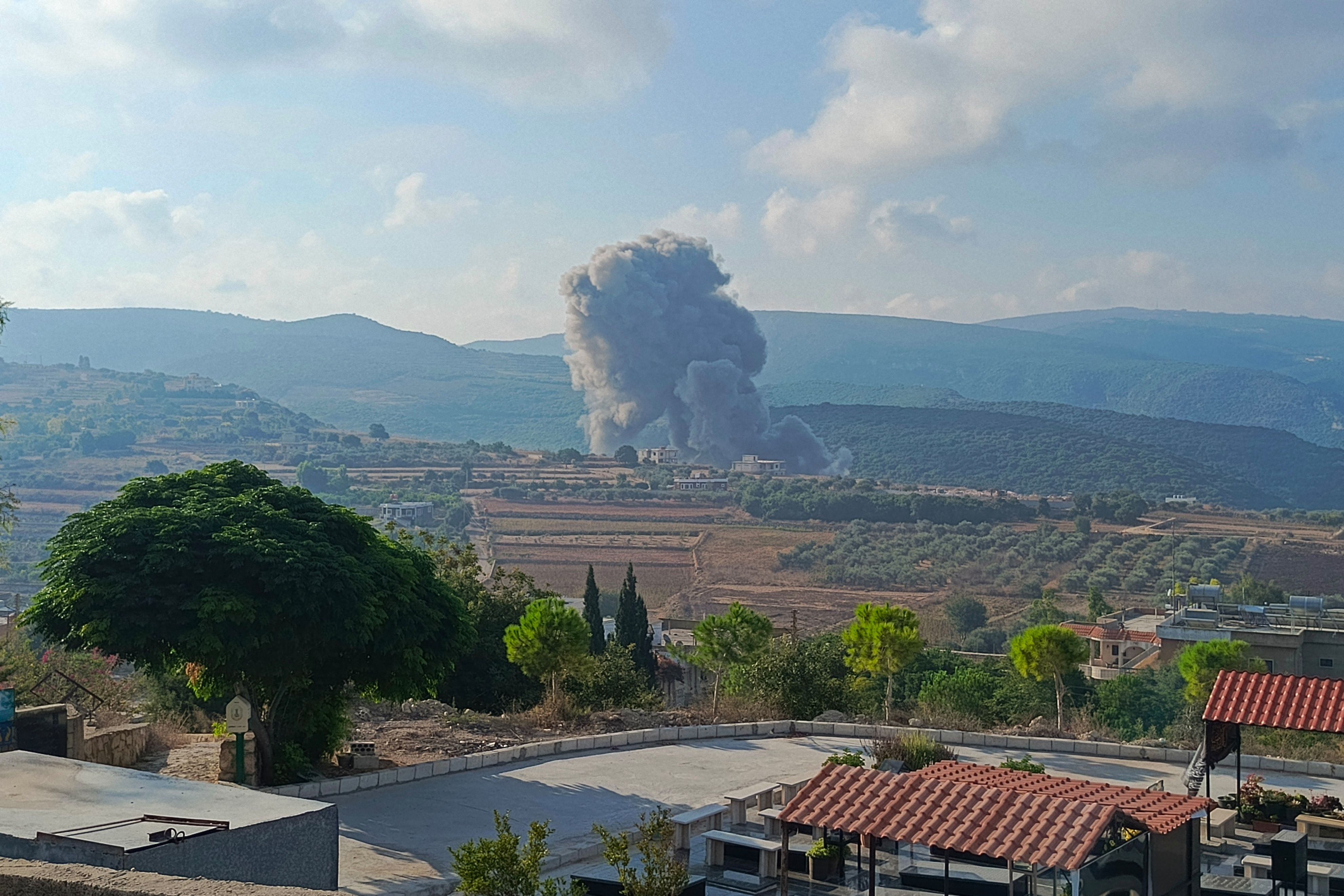 Smoke rises in the distance in this wide shot of a landscape with trees and homes