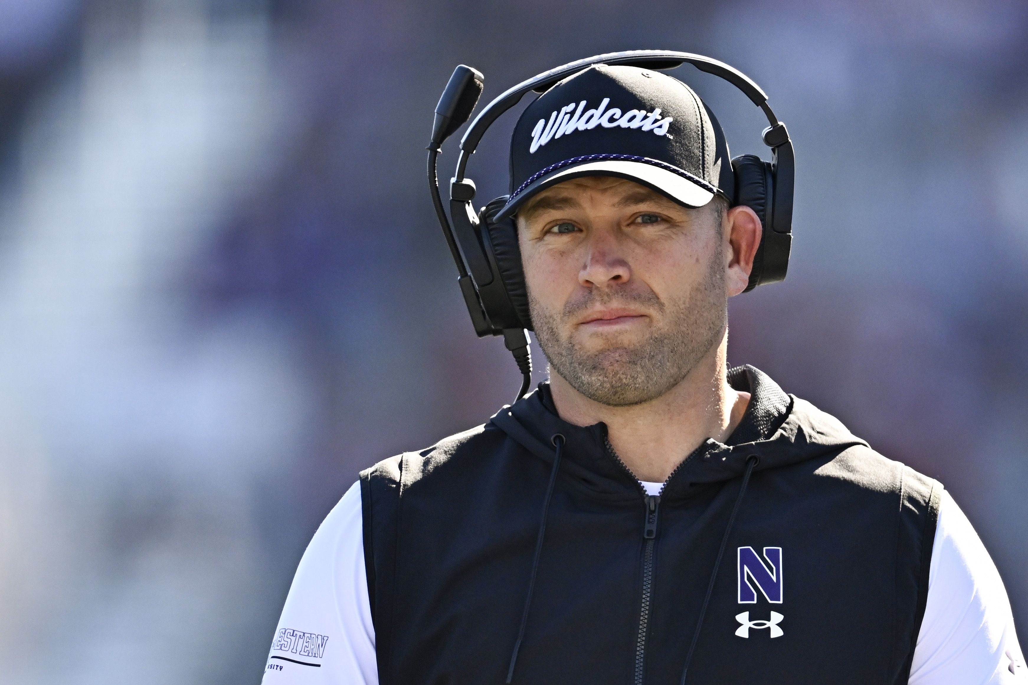 Man wearing black Wildcats cap and black sports vest with Northwestern logo, using headset microphone, looks focused against a blurred background.