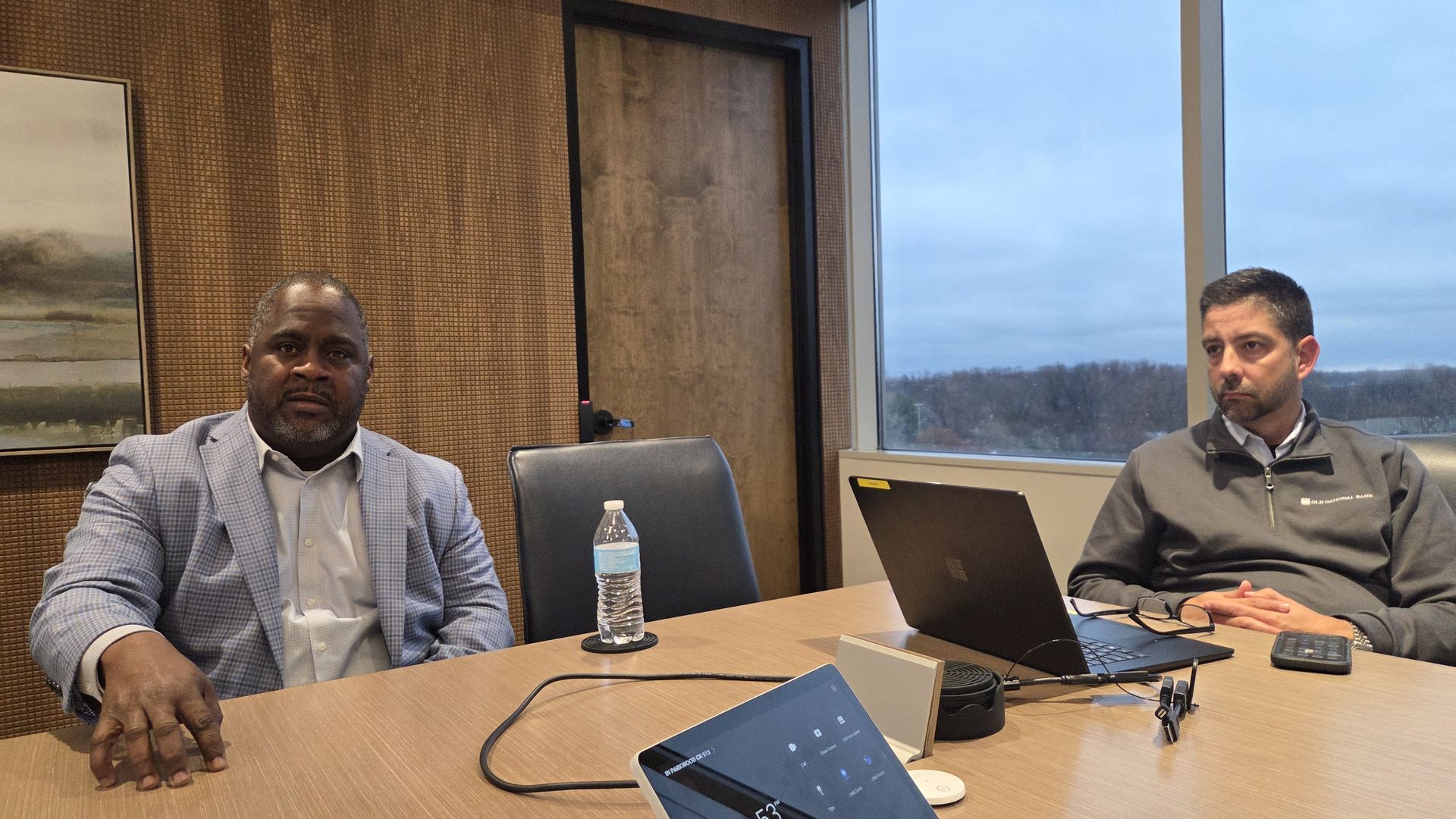 Two men sit in a modern office meeting room with wood-paneled walls and large window showing trees. One in a light blue checkered blazer, the other in a gray pullover with laptop and water bottle on table.