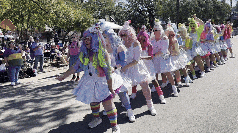 Image shows women in brightly colored wigs and aprons dancing in a parade.