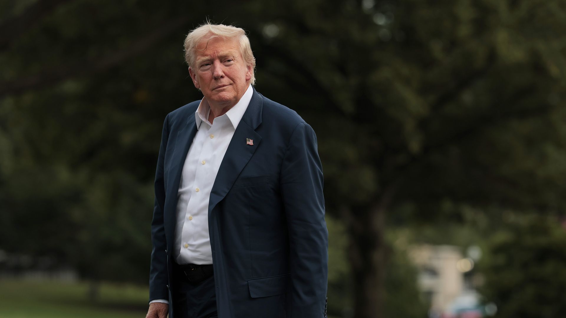 Trump walks across the South Lawn of the White House in a navy suit and a white collared shirt.