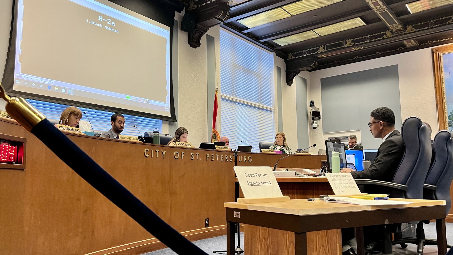 City council meeting in St. Petersburg with members seated behind a wooden panel labeled CITY OF ST. PETERSBURG, a large screen showing "H-2a 1. Brandi Gabbard," and an open forum sign on a table.